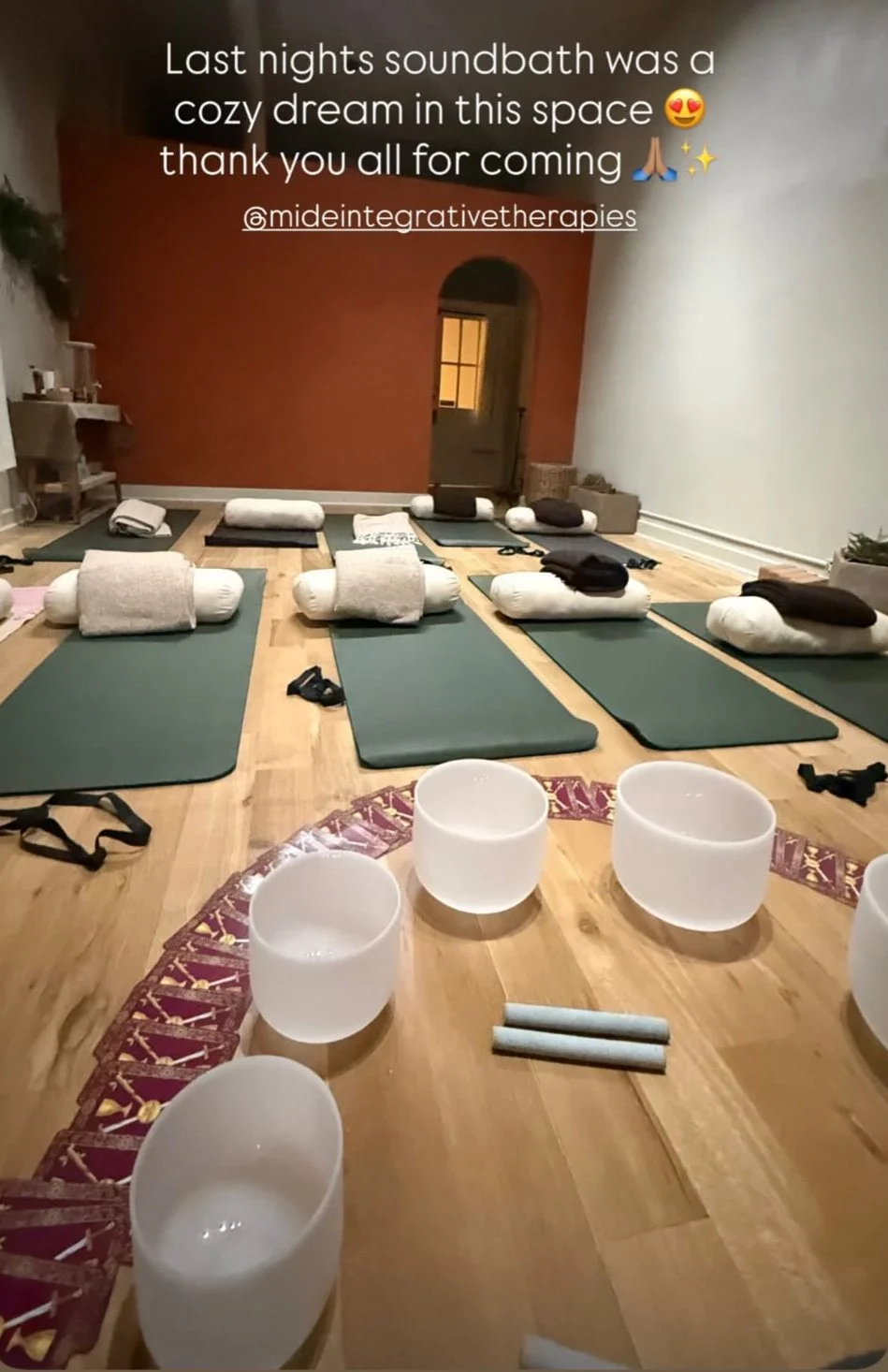 A cozy yoga or sound bath studio with yoga mats, bolsters, and blankets arranged on a wooden floor. There are crystal singing bowls in the foreground, and the back wall is painted a warm red. The space looks inviting for relaxation and meditation.