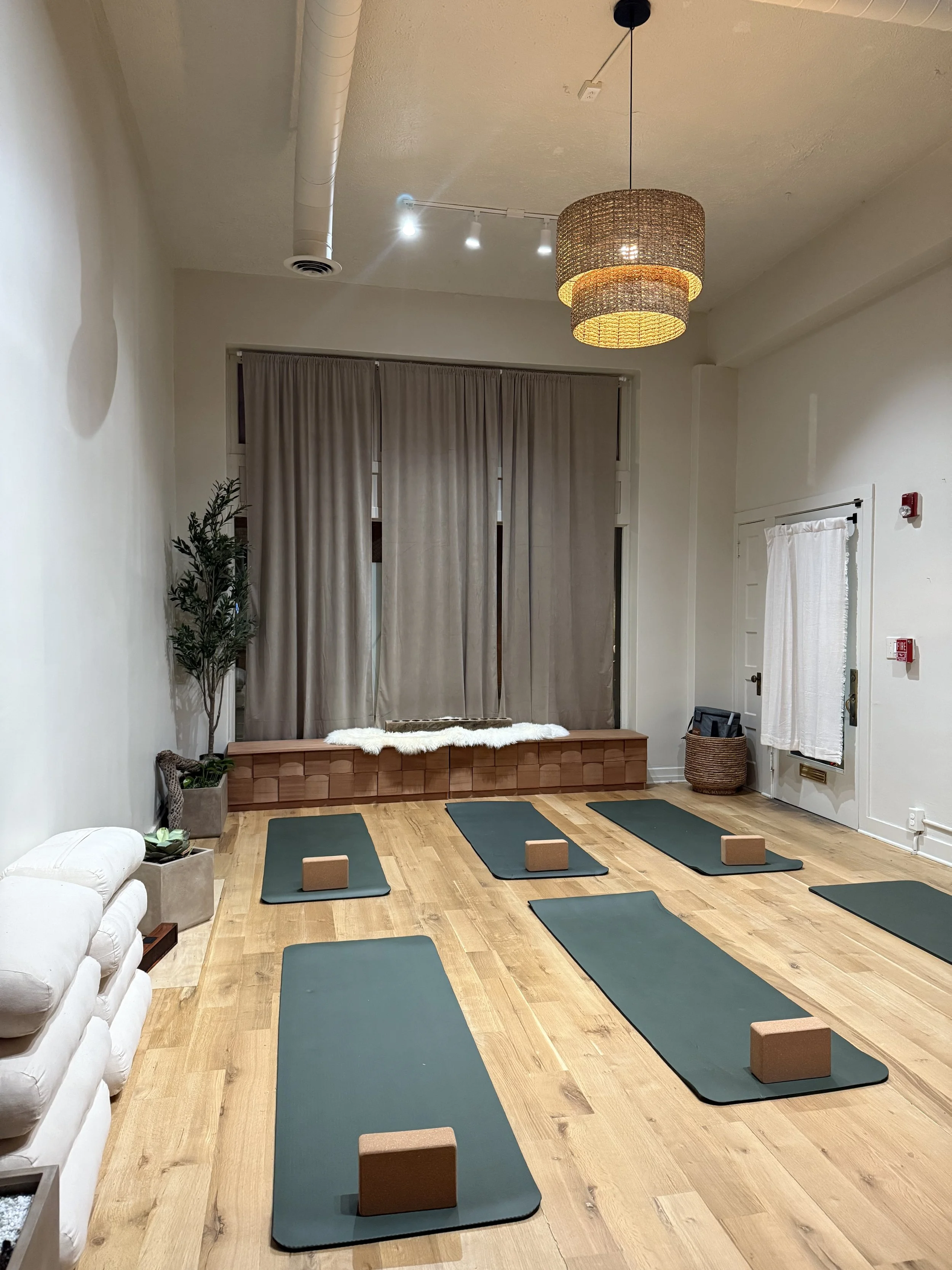 Yoga or meditation class setup in a studio with six yoga mats on a wooden floor, each with a cork yoga block, a white couch on the left, potted plants, closed beige curtains, and a woven pendant light hanging from the ceiling.