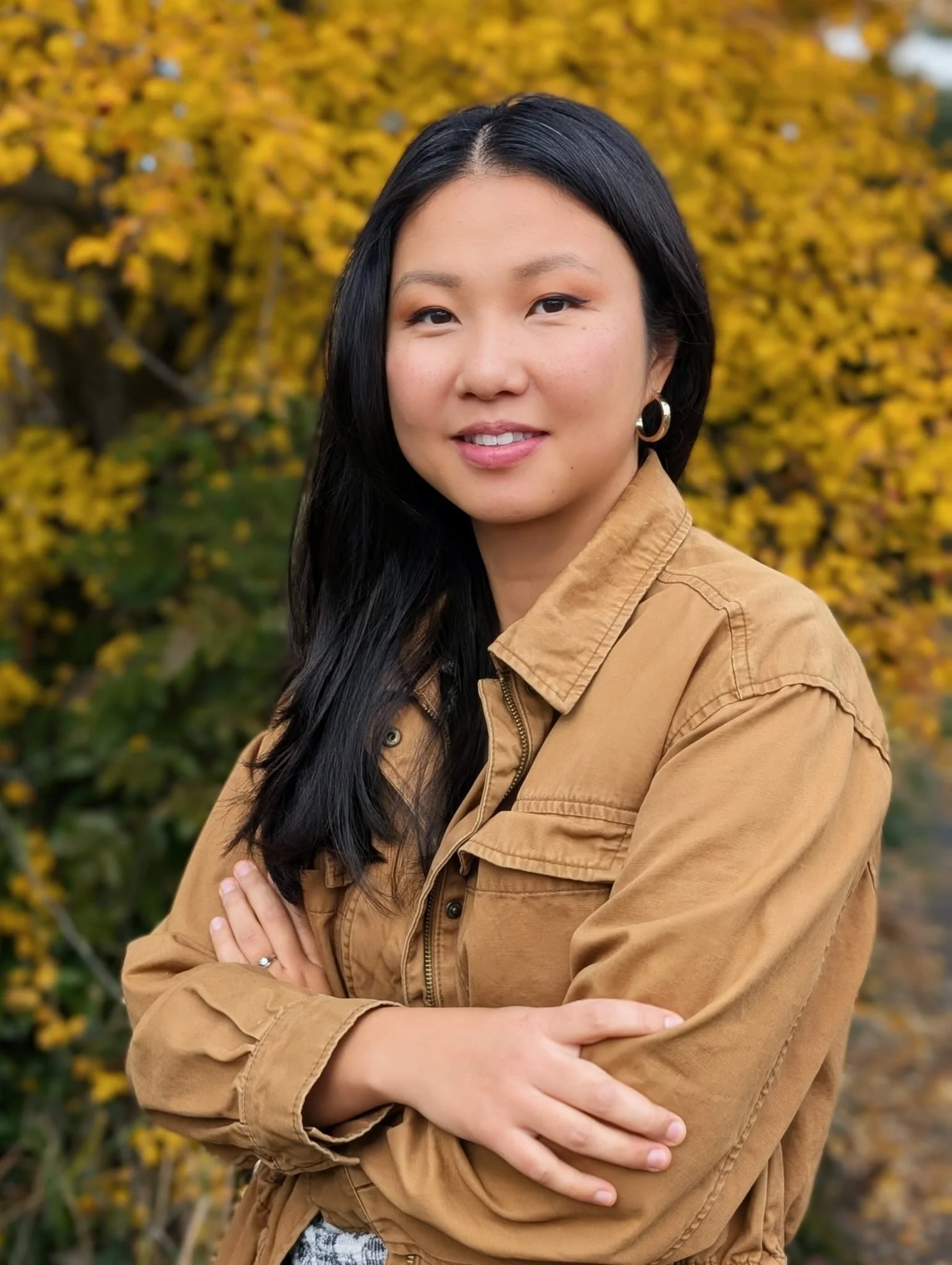 A woman with long black hair and hoop earrings posing outdoors with her arms crossed, standing in front of a tree with yellow autumn leaves.