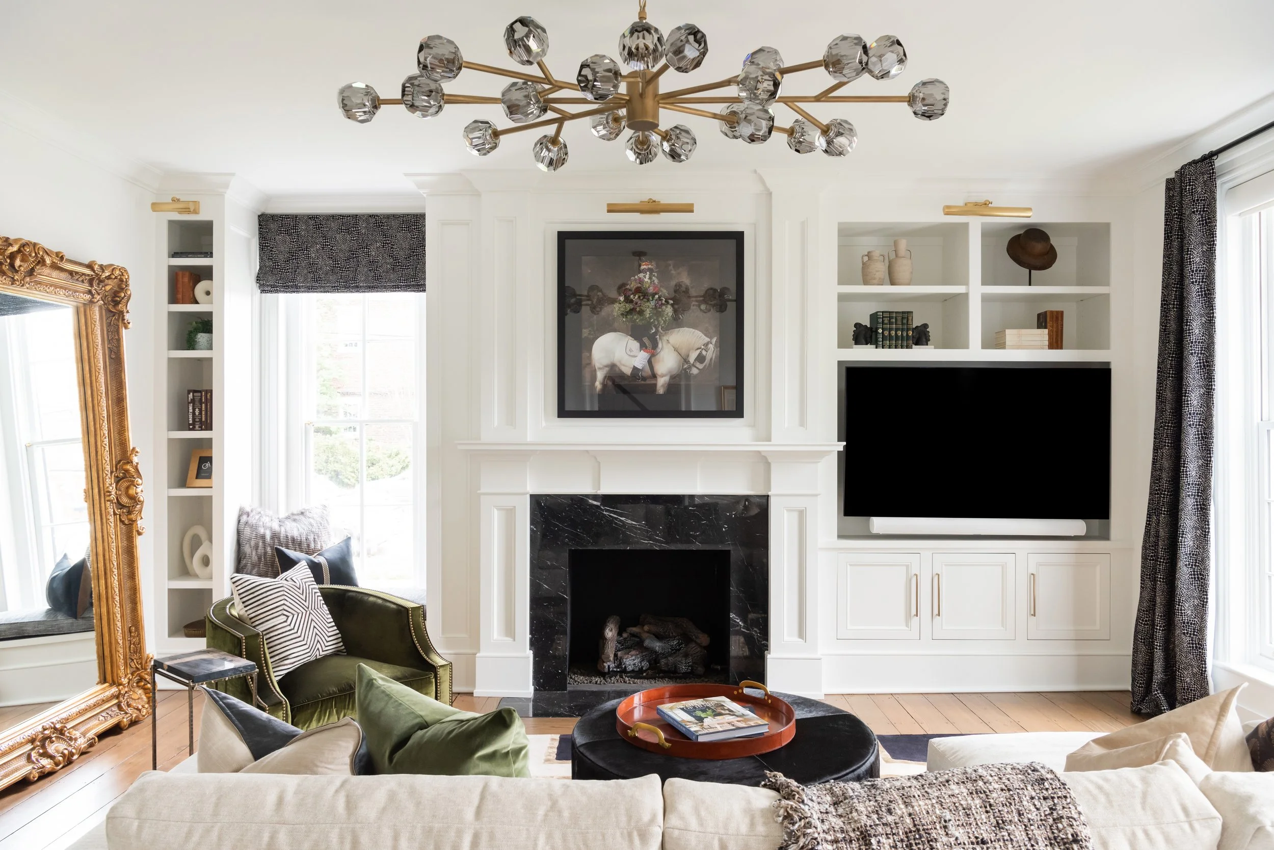 Living room with white built-in shelves, black marble fireplace, large TV, green velvet armchair with pillows, black ottoman with tray, gold mirror, window with patterned black curtains, and modern chandelier with gold and glass accents.
