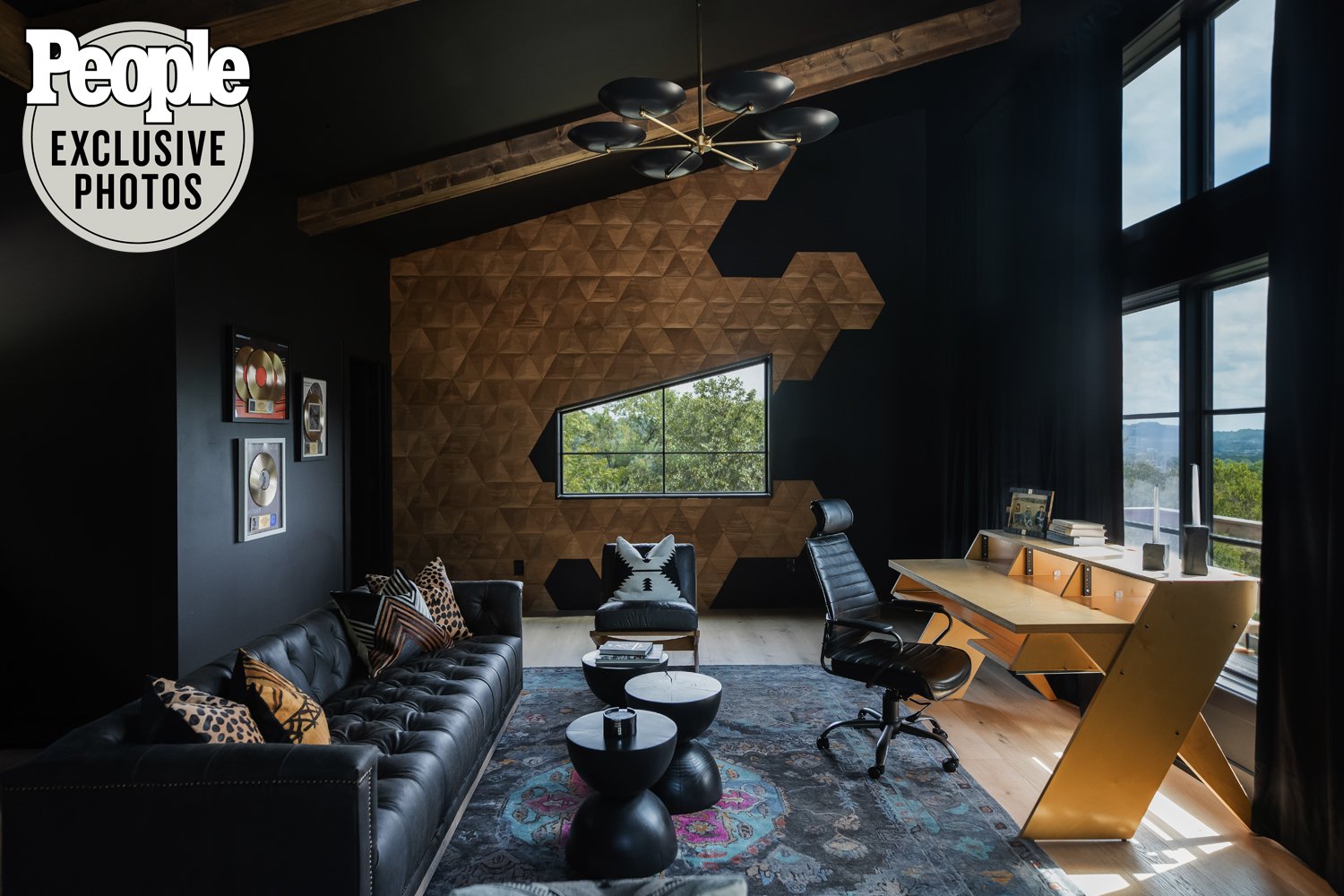 Modern home office with black leather sofa and patterned pillows, wooden desk with black hardware, black office chair, large window with greenery view, framed record awards, geometric wood accent wall with irregular window, and black chandelier.