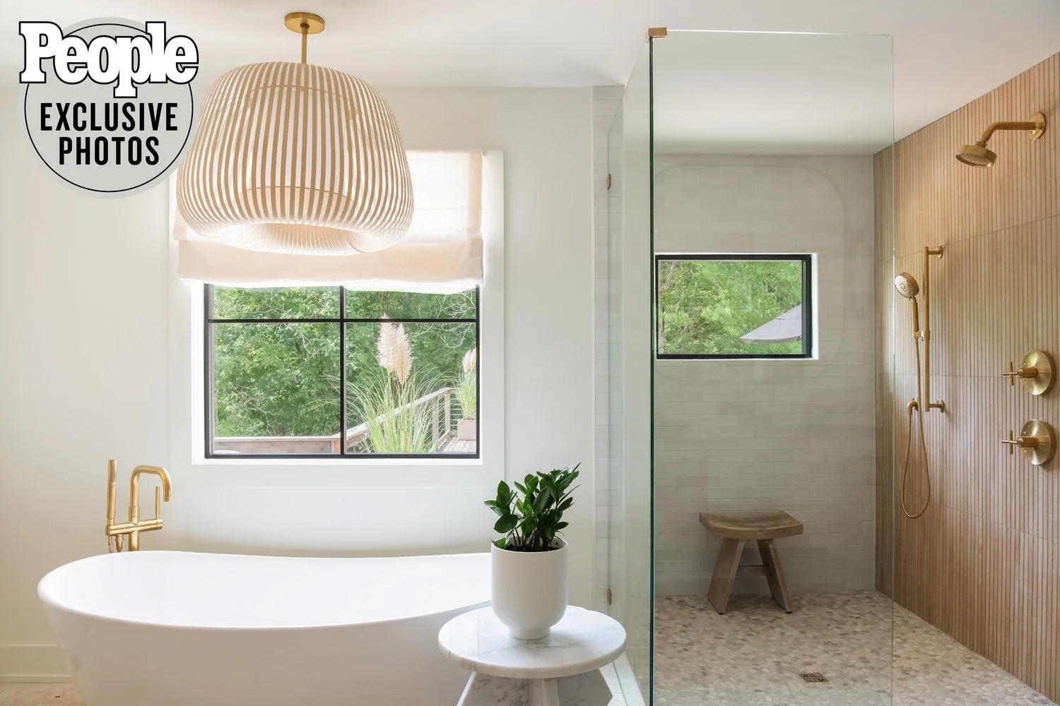 Modern bathroom with white freestanding bathtub, round marble side table with potted plant, large window with tree view, wooden bench in shower area, and brass ceiling light fixture.