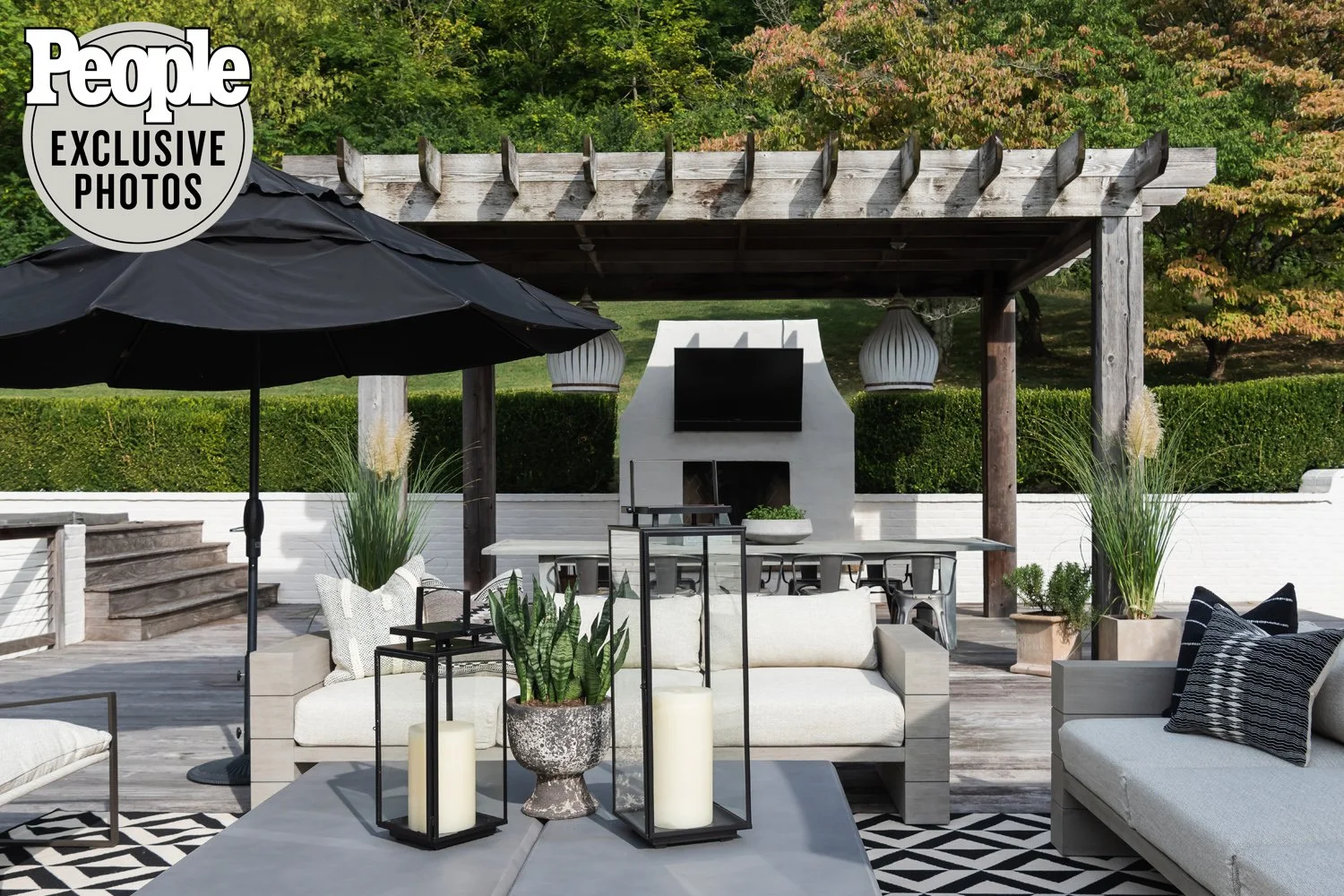 Outdoor patio with white furniture, black umbrella, lanterns, potted plants, fireplace, and flat-screen TV under wooden pergola, surrounded by greenery and trees.