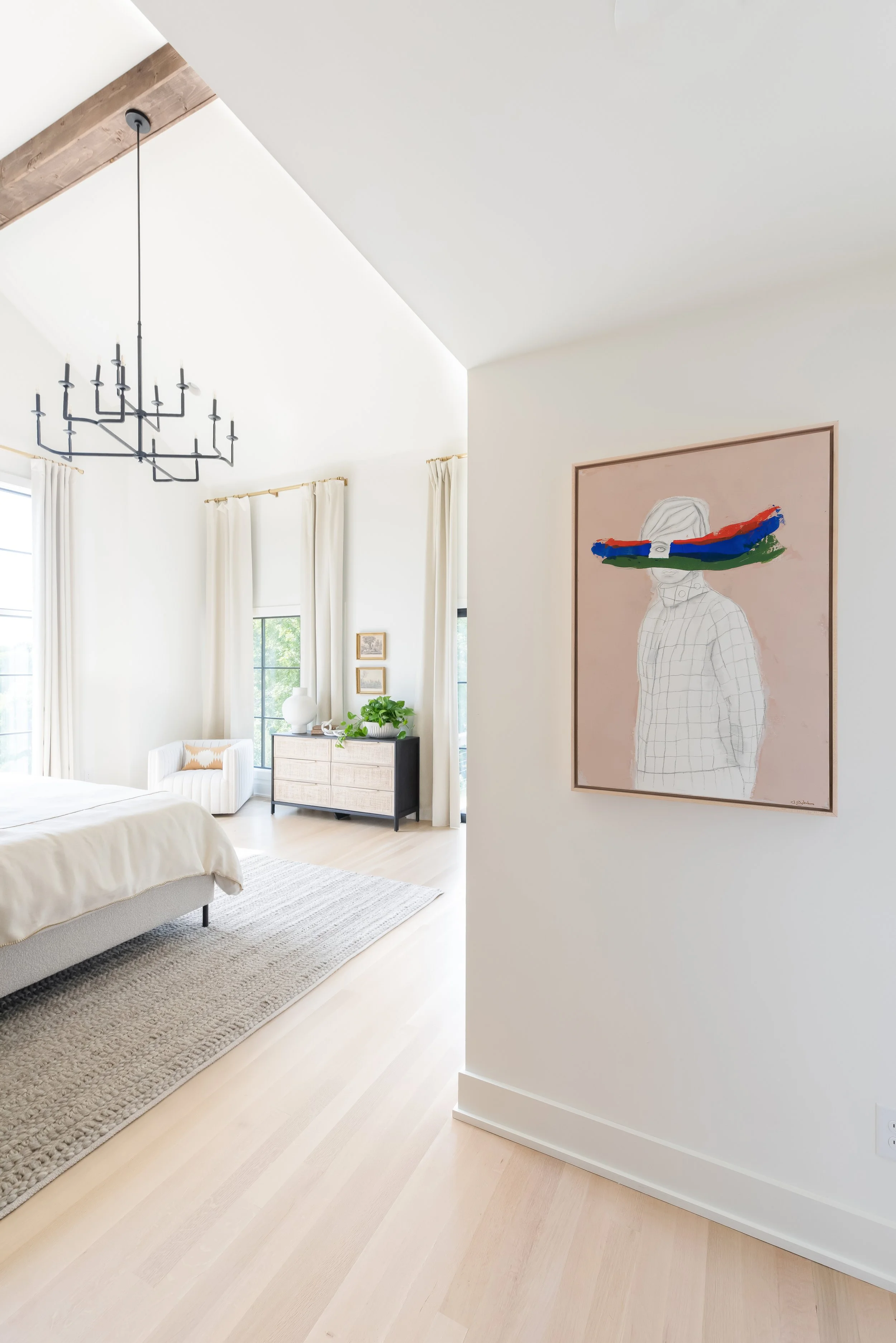 Bright bedroom with white walls, exposed wood beam ceiling, large black chandelier, and beige curtains, featuring a light-colored bed, window-side chair, dresser with plant and decor, and abstract artwork with colorful striped eyes.