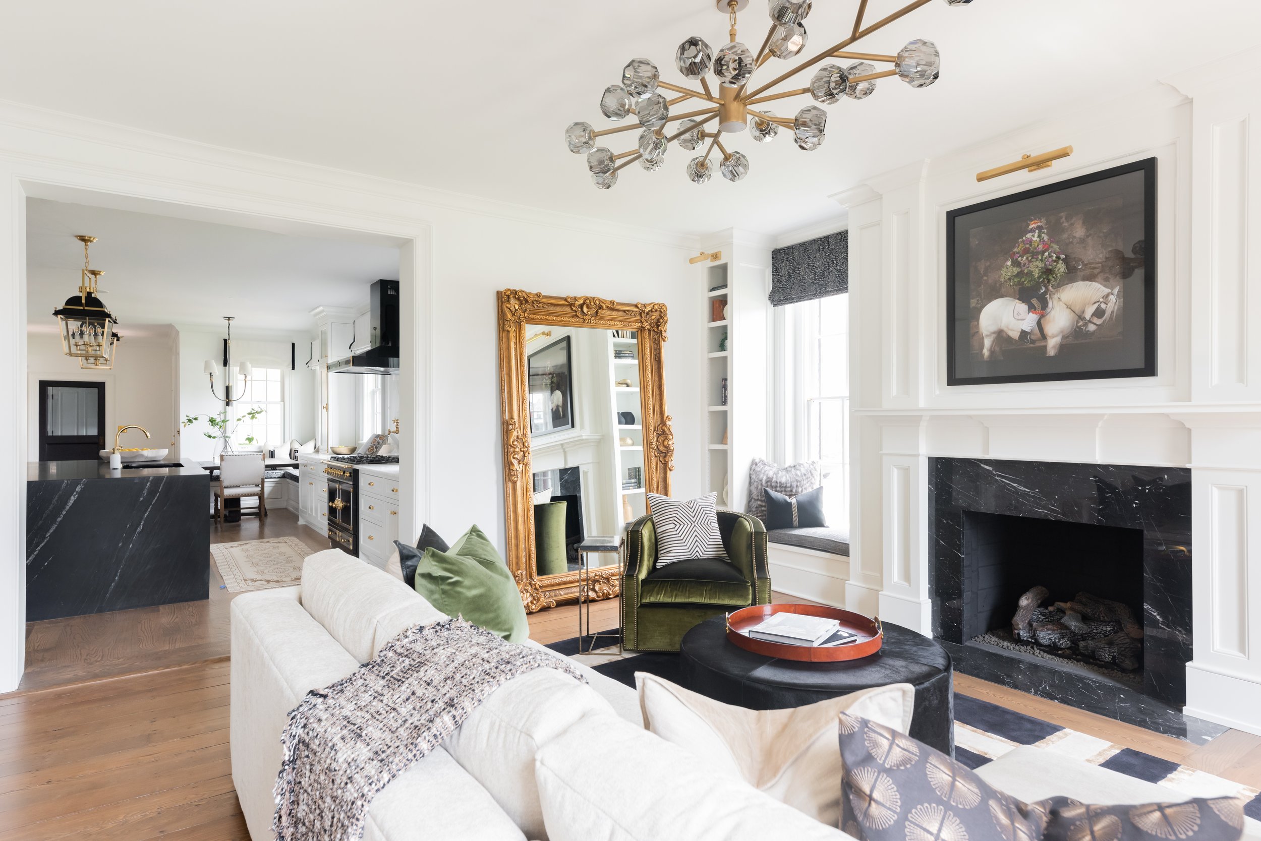 Living room with white walls, black marble fireplace, framed horse artwork with floral arrangement, white sofa, green armchair, black ottoman, gold-framed mirror, modern gold chandelier, and window seat with pillows.