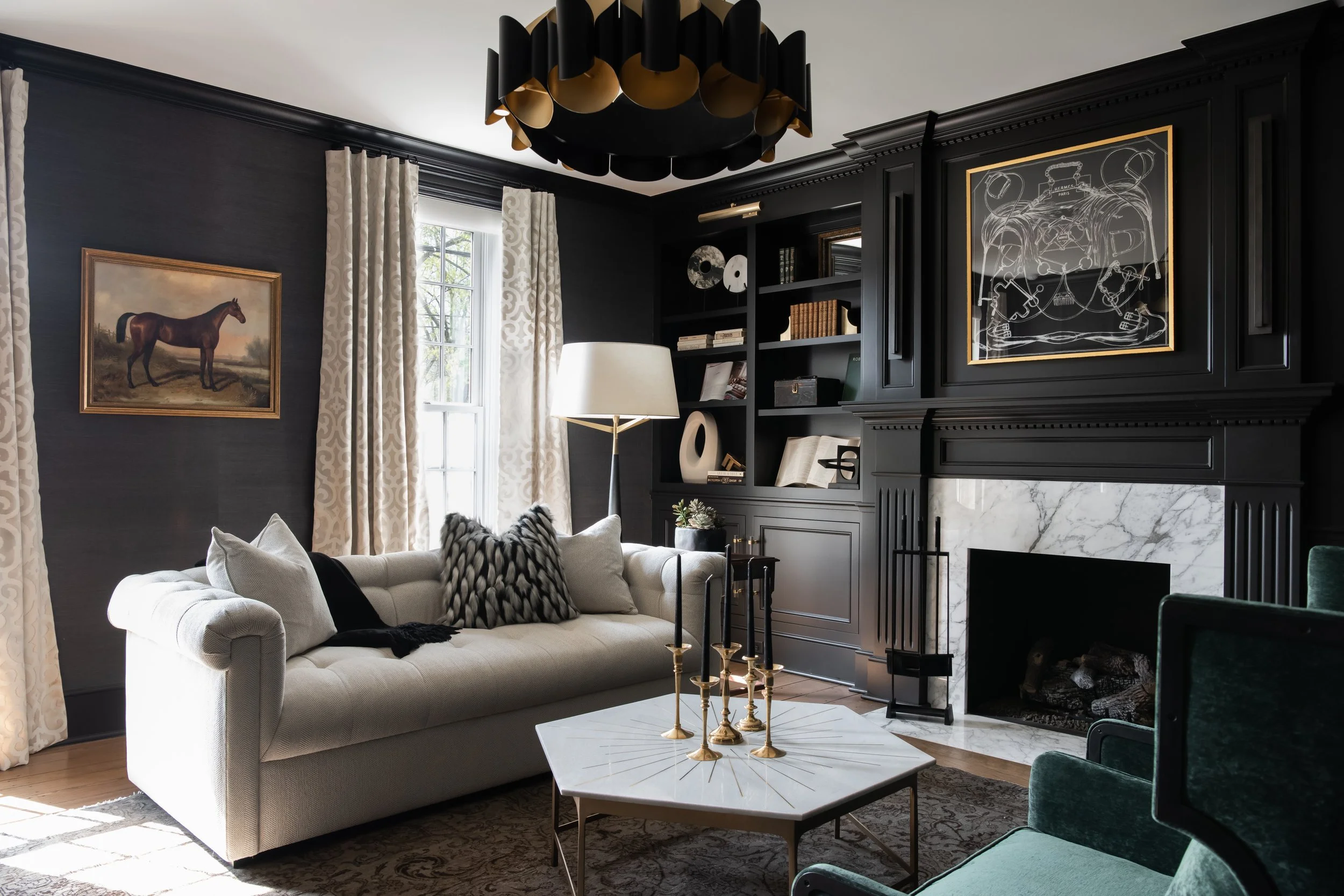 Living room with black paneled walls, white fireplace with marble surround, cream sofa with pillows, black bookshelf, window with patterned curtains, black and gold chandelier, and white marble coffee table with gold candlesticks.