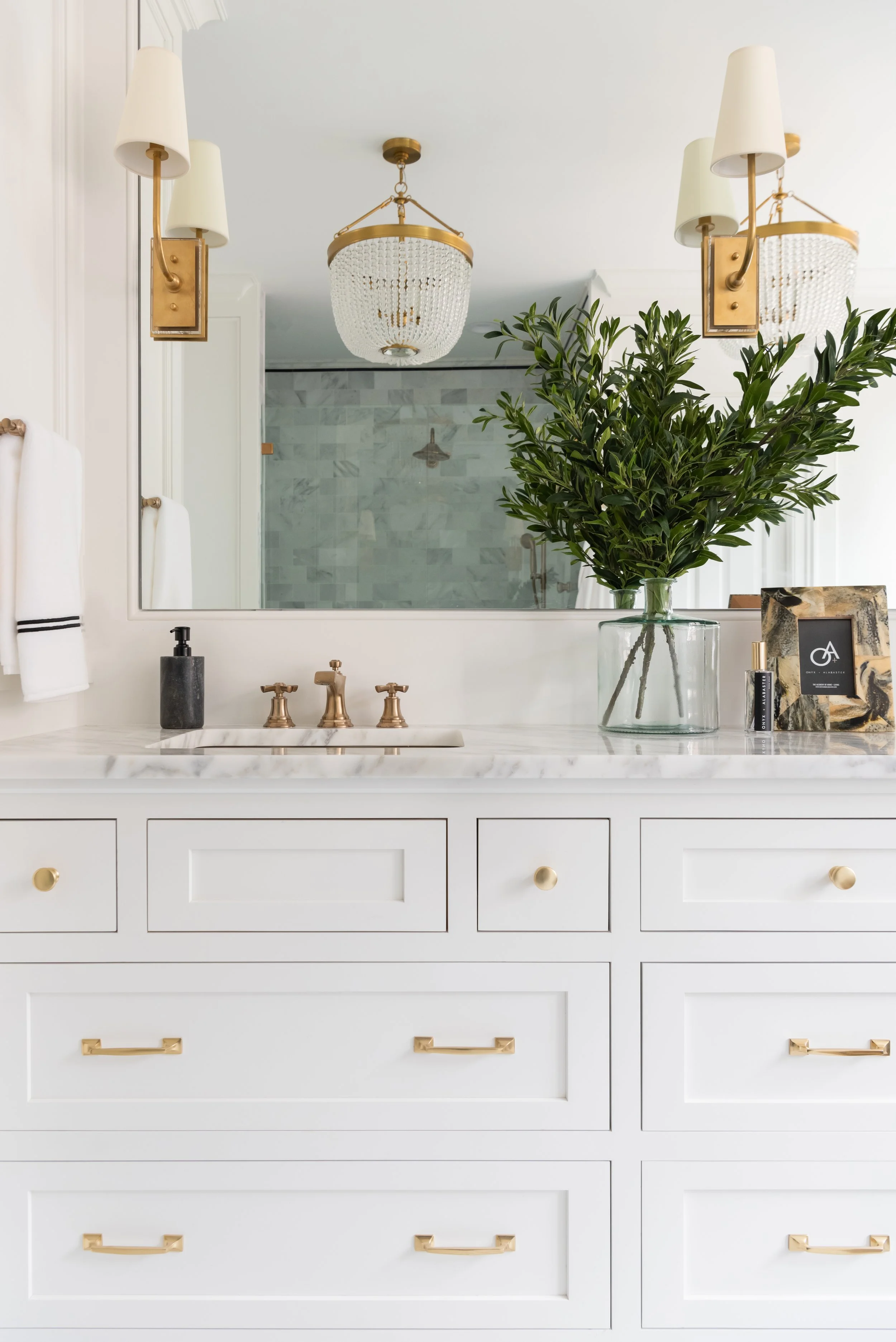 White bathroom vanity with gold hardware, large mirror, vase with greenery, and soap dispenser, with chandelier and shower visible in reflection.