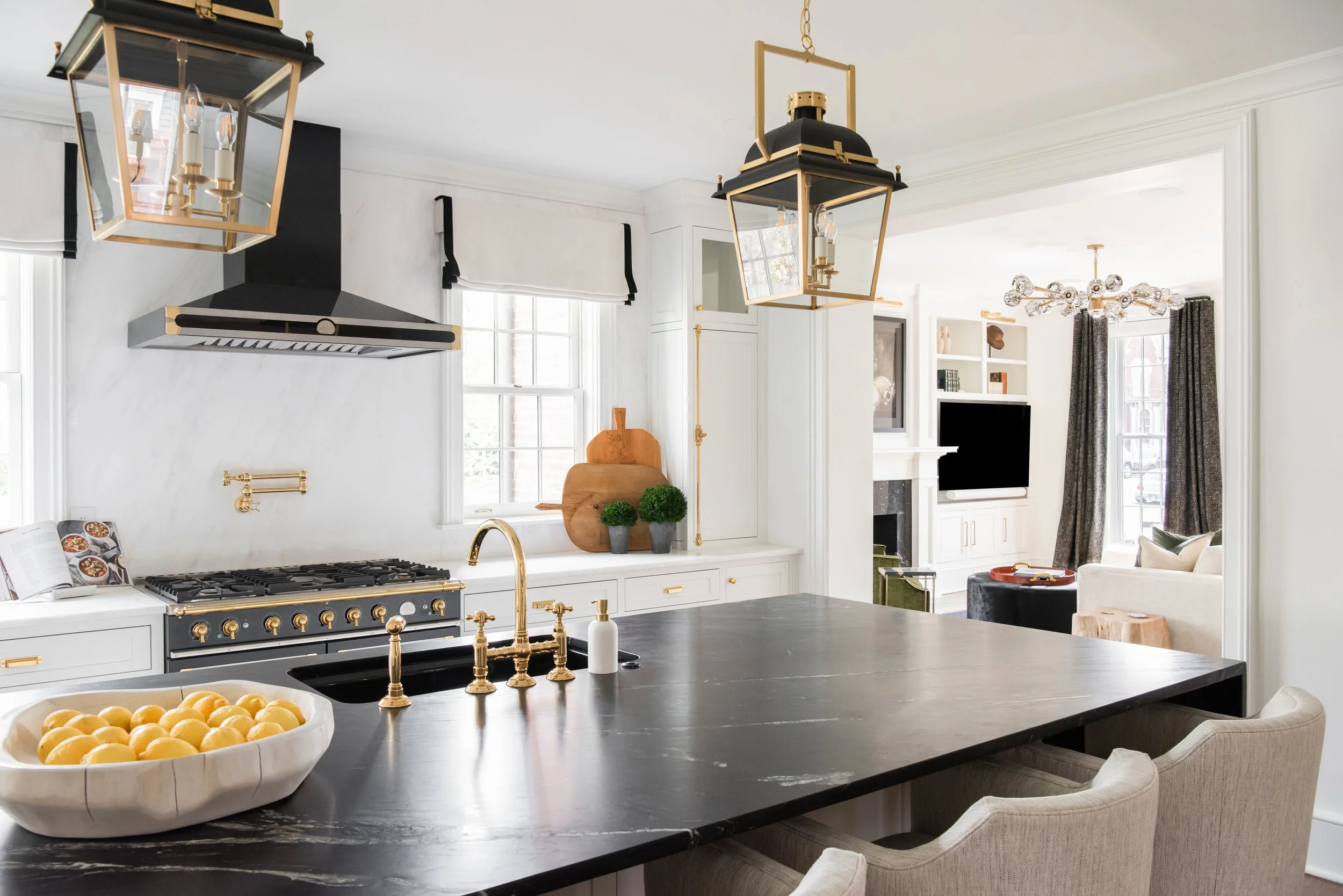 Modern kitchen with black island, gold fixtures, white cabinets, open layout to living room with TV, sofa, and large windows.