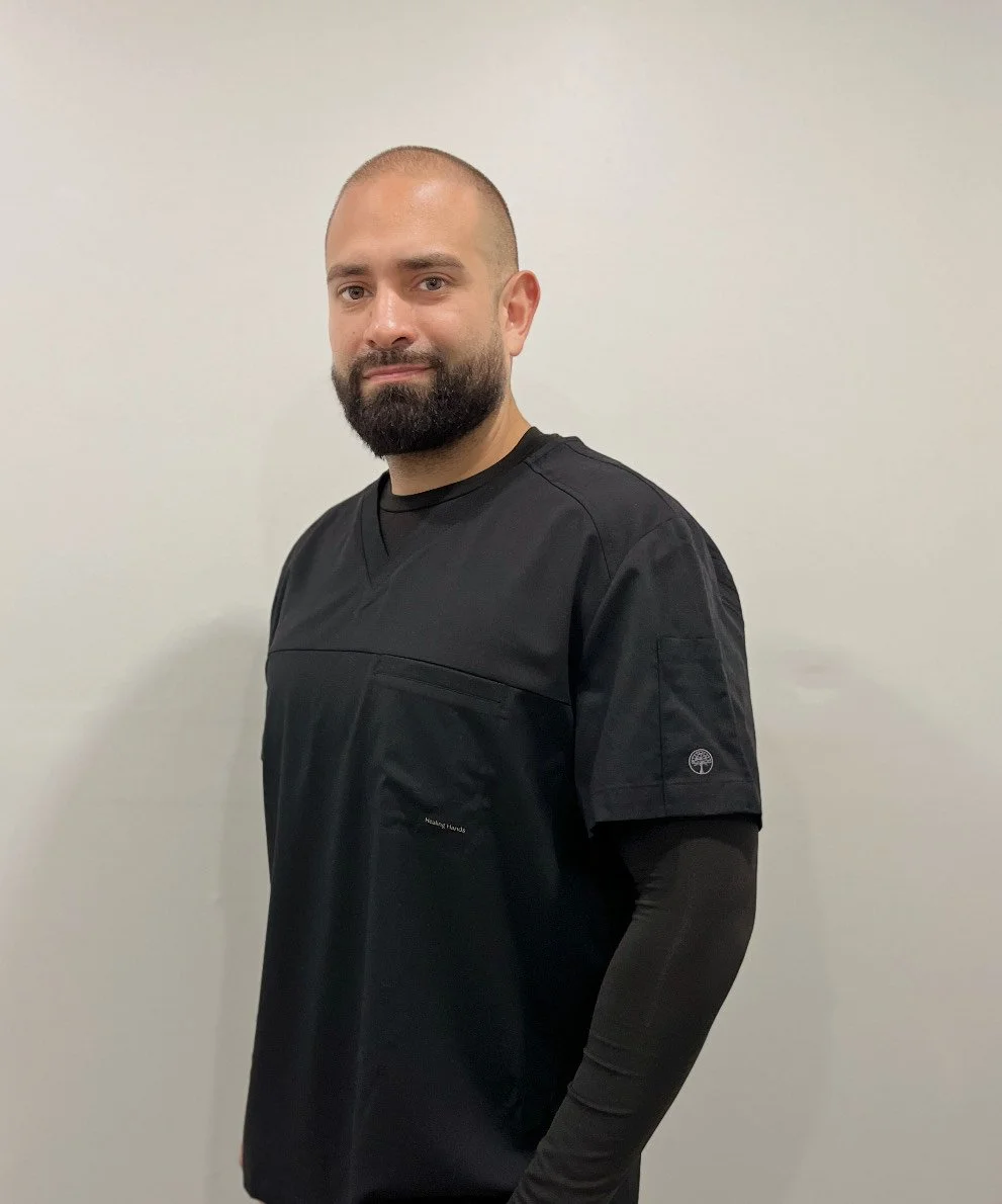 A man with a beard and short hair wearing black medical scrubs standing against a plain white wall.