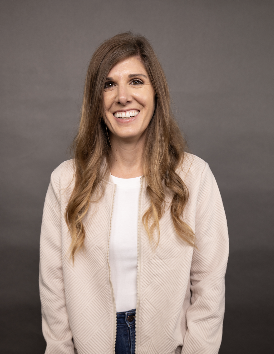 Portrait of a smiling woman with long, wavy light brown hair, wearing a light-colored zip-up jacket over a white shirt, standing against a plain gray background.