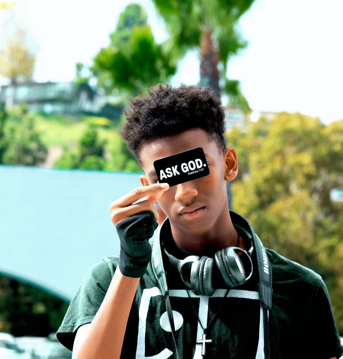 A young man with dark curly hair holds a black sign over his eye that says "ASK GOD" with a scripture reference, outdoors with green trees and a blue sky in the background.