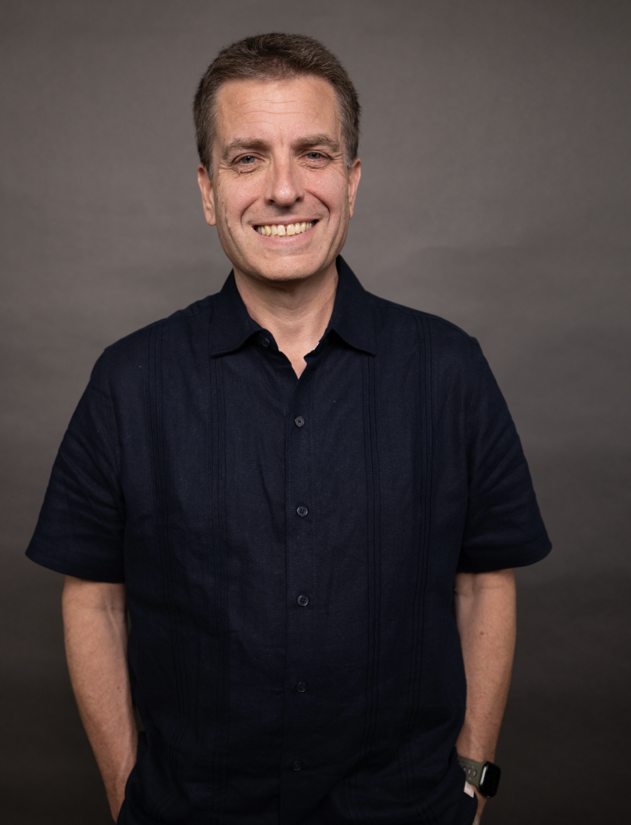 A middle-aged man with short brown hair, wearing a navy blue short-sleeve button-up shirt, smiling, standing against a plain gray background.