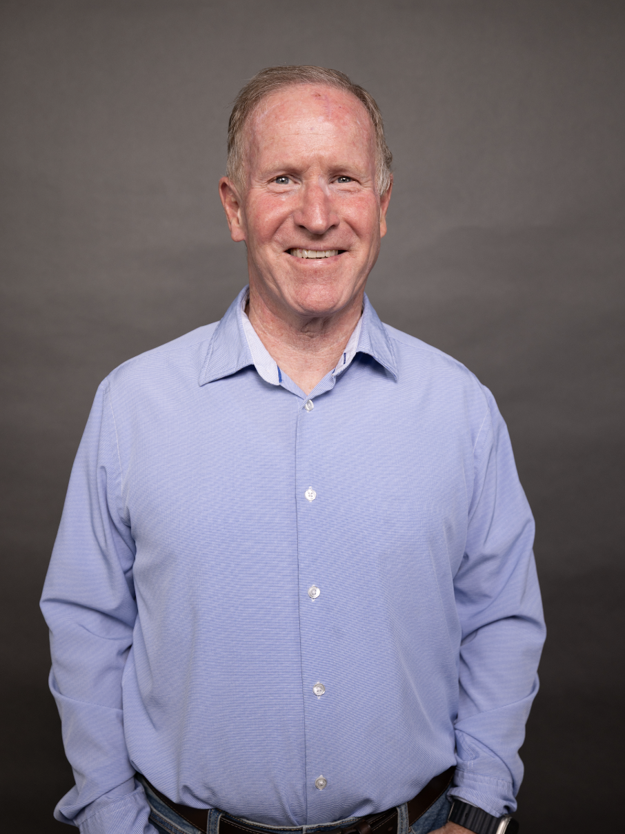 A smiling middle-aged man with short, light-colored hair wearing a light blue button-up shirt standing against a plain gray background.