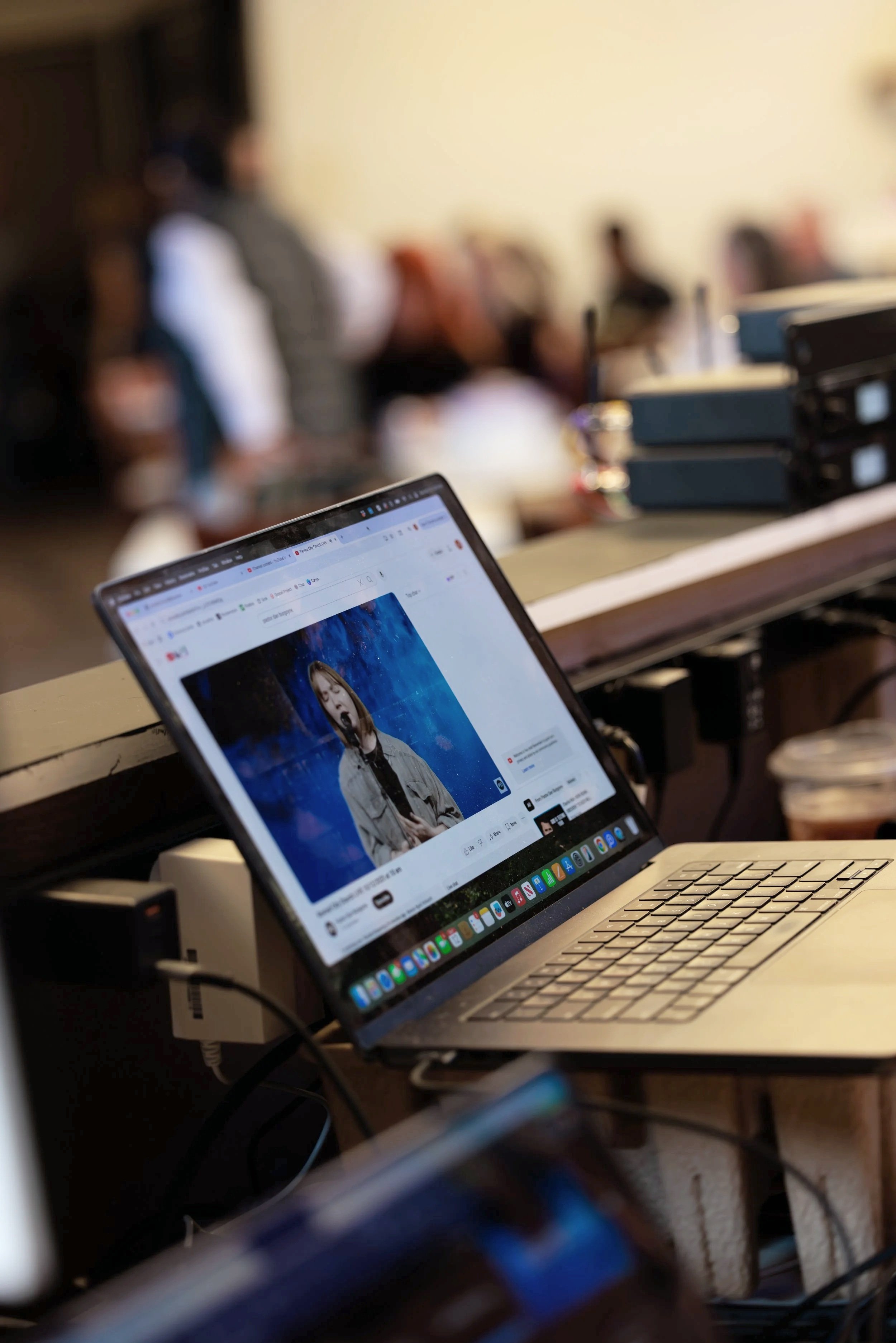 Laptop on a wooden table in a cafe shows a paused YouTube video of a female singer performing against a blue background with blurred people in the background.