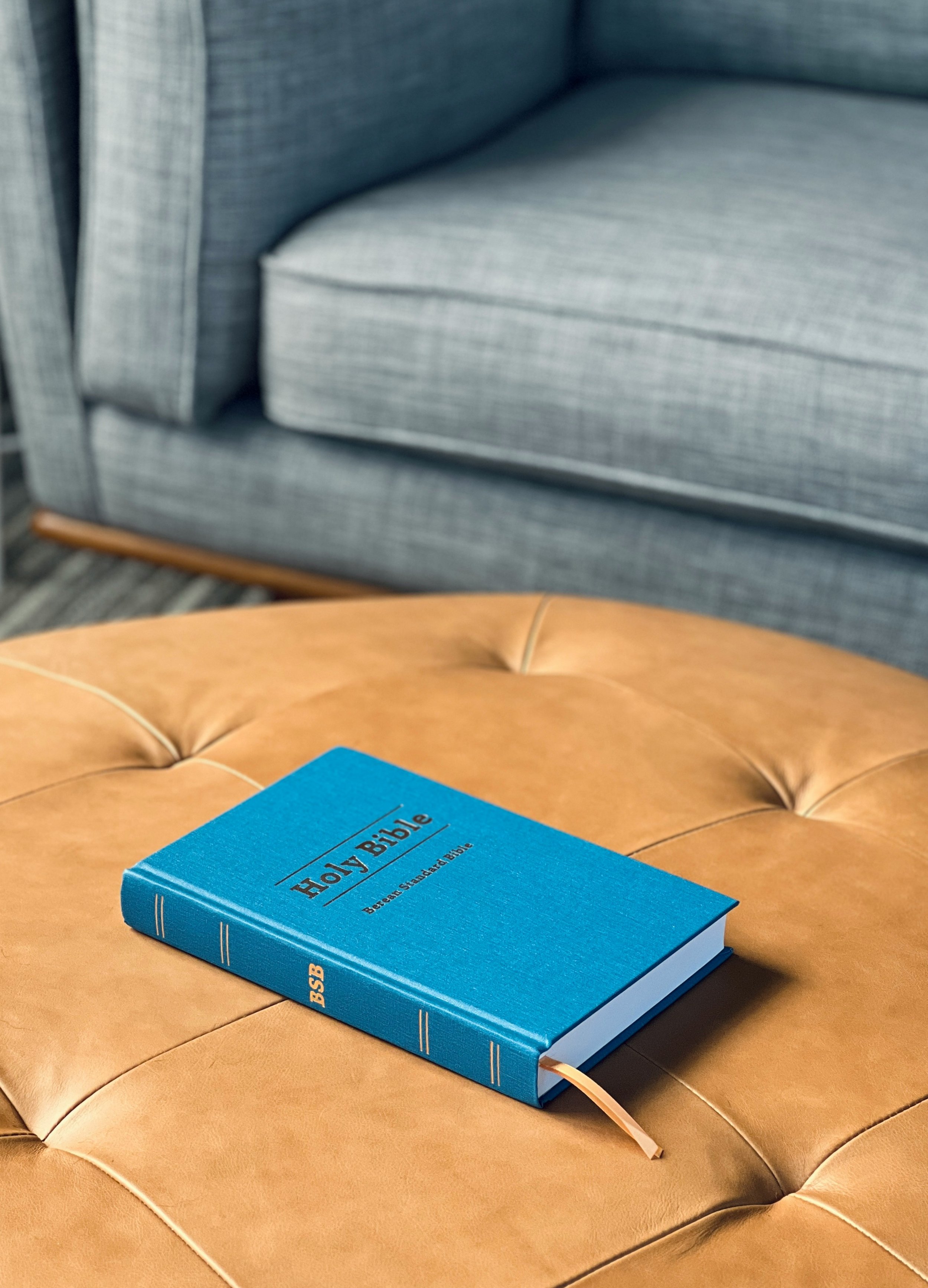 A blue hardcover Holy Bible resting on a tan tufted leather ottoman with a gray upholstered armchair in the background.