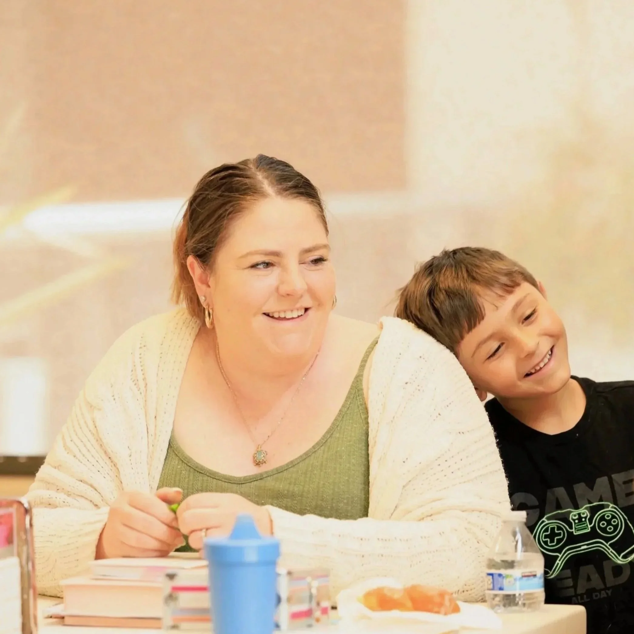 A woman and a young boy sitting at a table, smiling and enjoying each other's company. The woman has short brown hair, is wearing a green top with a white cardigan, and a necklace. The boy has brown hair and is wearing a black T-shirt.