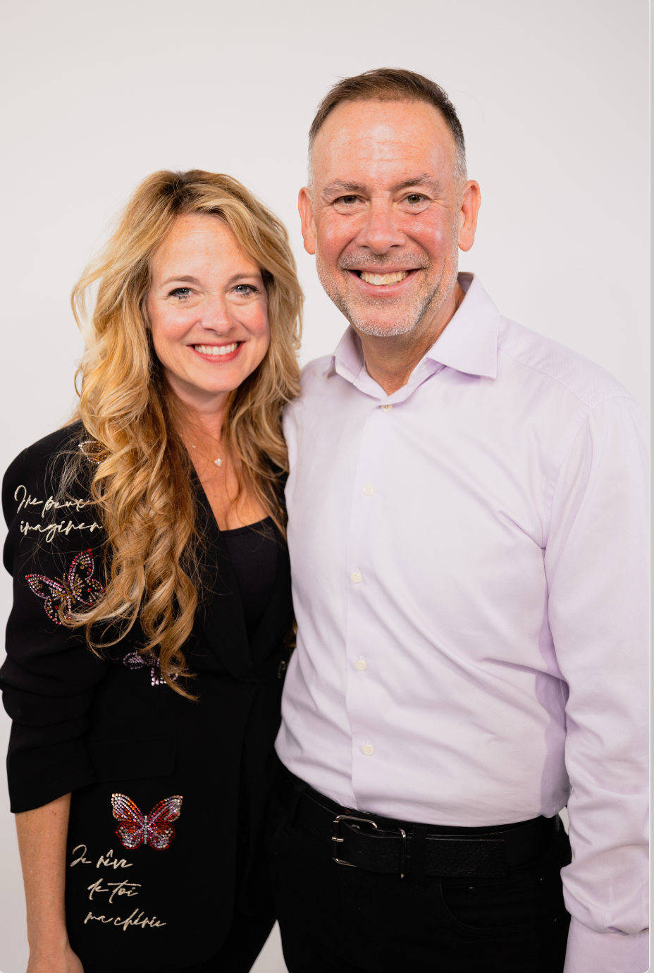 A smiling couple stands together, the woman with long blonde hair wearing a black blazer with embroidered butterflies and writing, and the man with short hair and a beard wearing a light pink shirt against a plain white background.