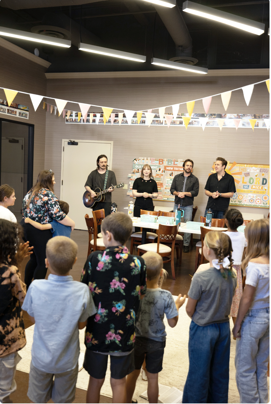 Children and adults gather in a room, listening to a musical performance by four musicians standing at the front. One musician is playing a guitar. The room is decorated with colorful triangular pennant banners and photos on the walls, and a biblical