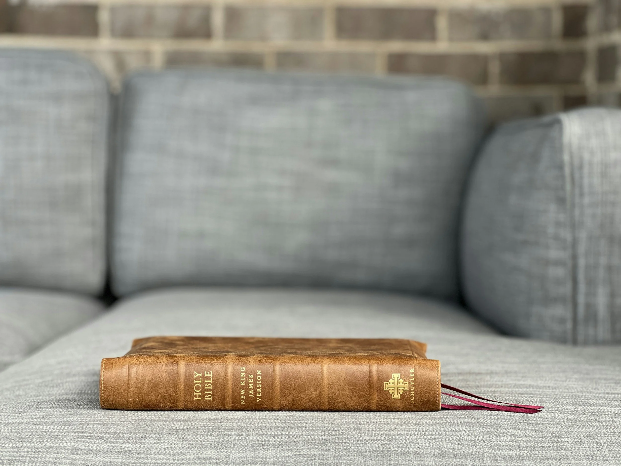 Brown leather-bound Holy Bible resting on a light gray fabric sofa with a brick wall background.