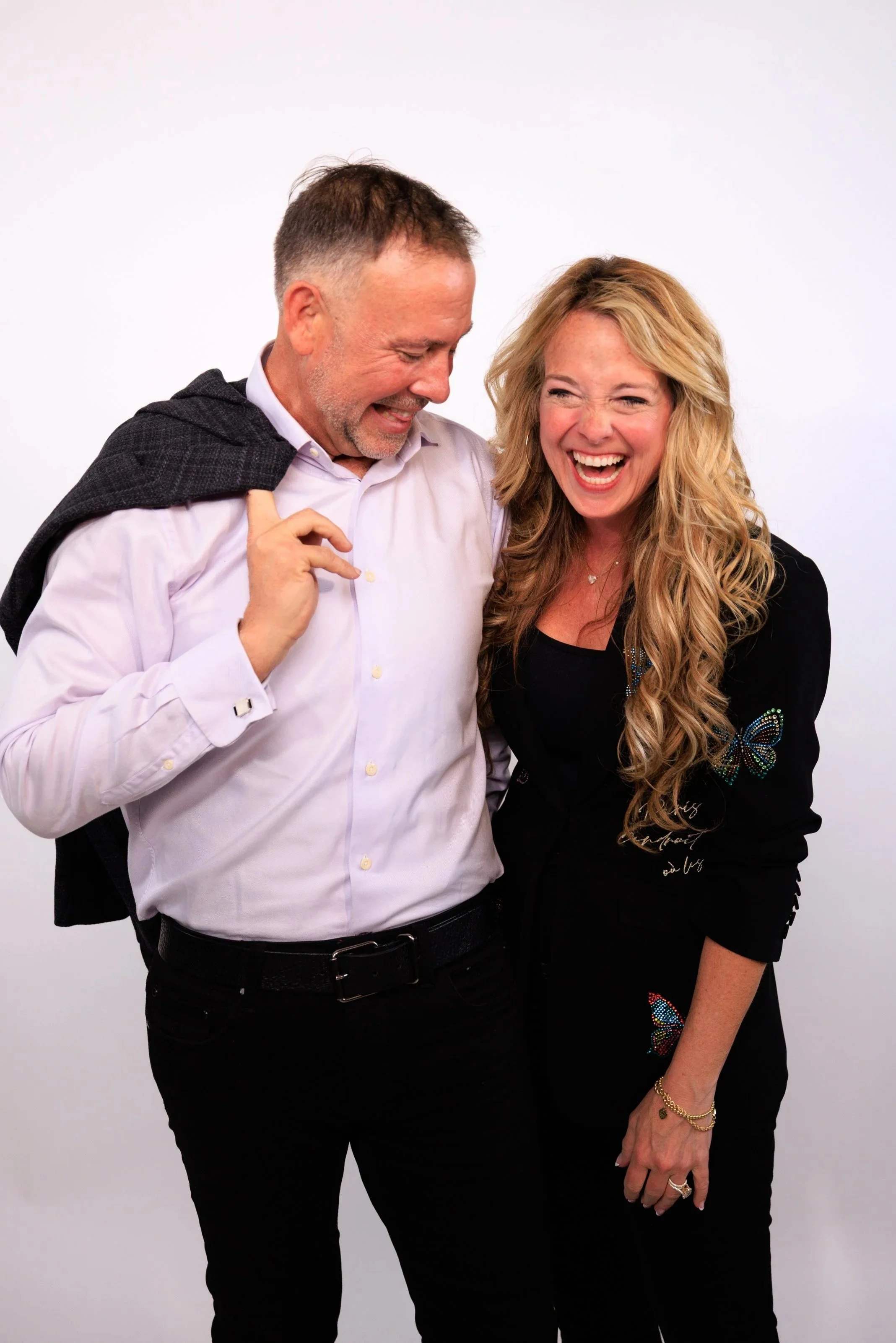 Two happy people, a man and a woman, laughing together against a plain white background. The man is wearing a white shirt and has a jacket draped over his shoulder, while the woman is dressed in a black top with butterfly designs and jewelry.