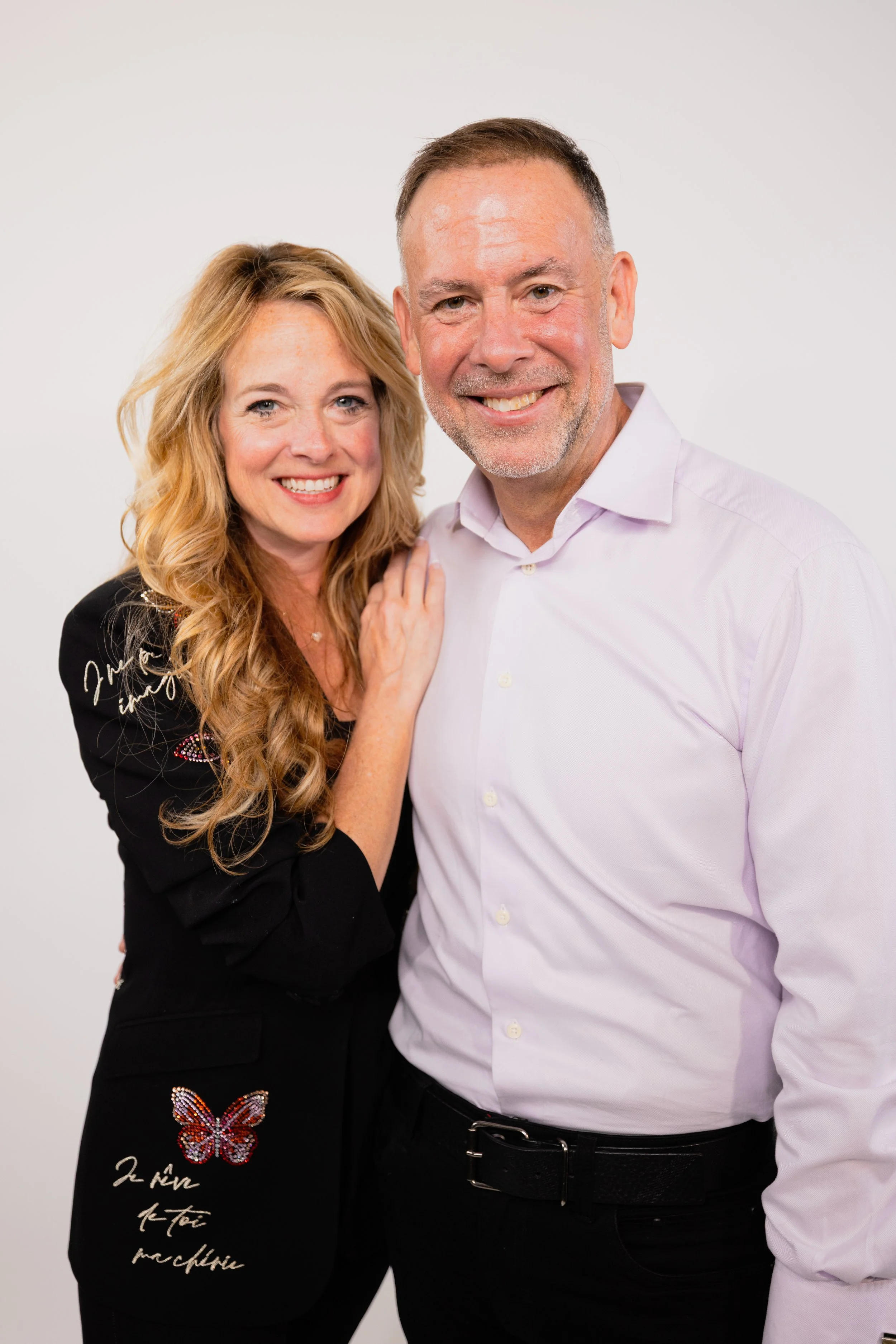 A smiling middle-aged couple standing close together against a plain white background, the woman has blonde curly hair and is wearing a black jacket decorated with a butterfly and text, and the man has short brown hair and a beard, wearing a white dr
