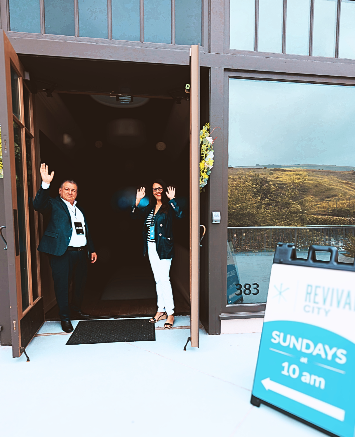 Two people standing at the entrance of a building waving. One man and one woman, dressed in business casual attire. A sign outside indicates Sunday hours at Revival City, starting at 10 am, with an arrow pointing left.