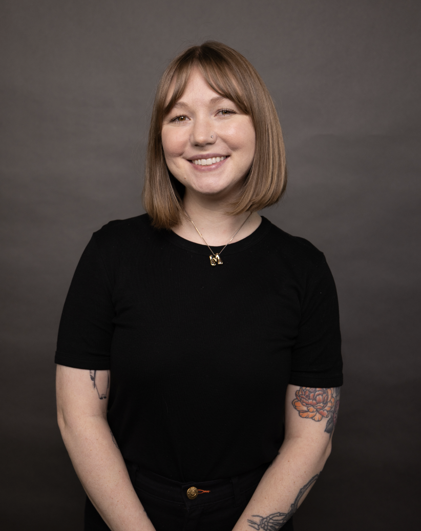 A young woman with shoulder-length light brown hair, wearing a black top, smiling, with tattoos on her arms and a nose piercing, posing against a gray background.