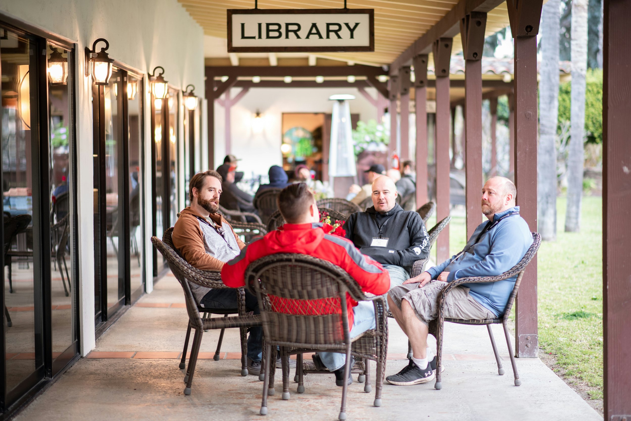 A group of five men sitting outside at a table on a covered patio, engaged in conversation, with a sign above that reads 'LIBRARY' and other people seated further back.