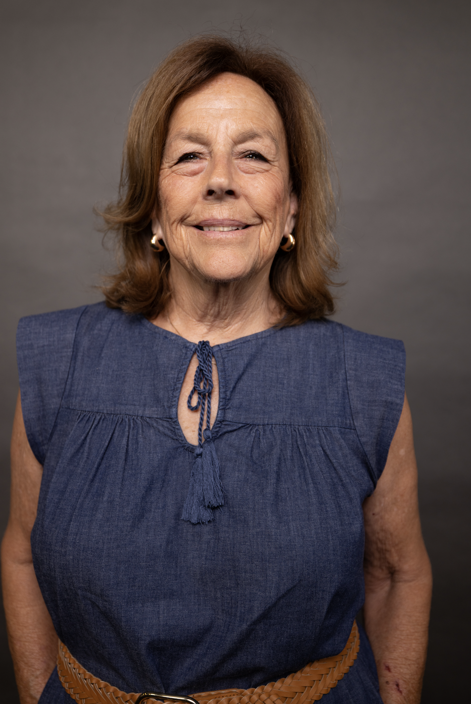 Smiling older woman with brown hair, wearing gold earrings, a blue sleeveless top with a tie detail, and a woven brown belt, standing against a gray background.