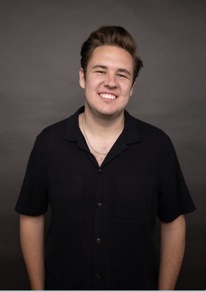 A young man with brown hair styled back, smiling and wearing a black short-sleeve button-up shirt and a silver necklace, standing against a dark gray background.