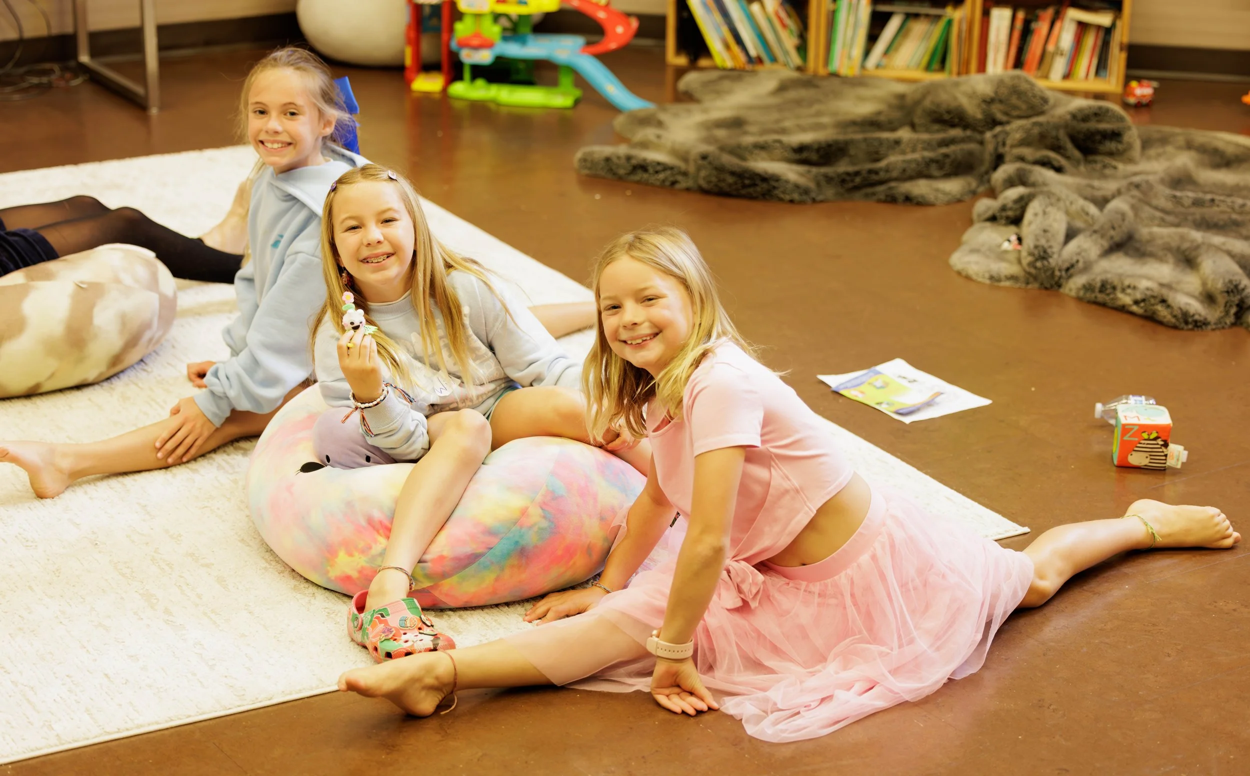 Three young girls with long hair sitting and lying on a cream-colored rug in a playroom, smiling at the camera. They are surrounded by toys, bookshelves, and large plush blankets.