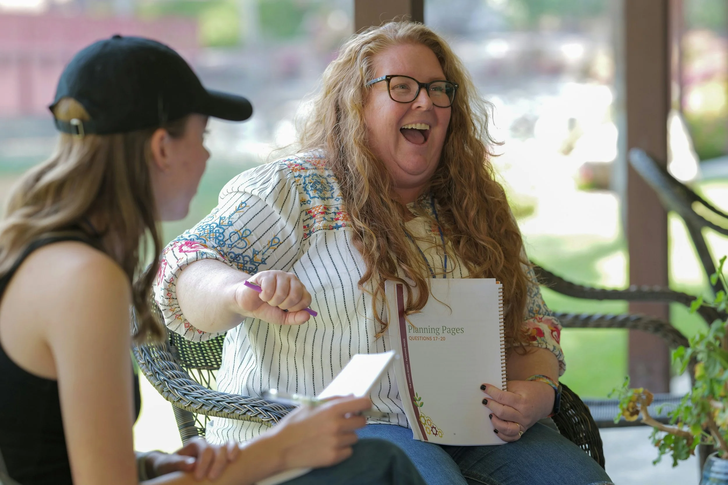 Two women sitting outdoors, one with long curly hair and glasses, holding a notebook and laughing, the other with long straight hair and a baseball cap, looking at a phone.