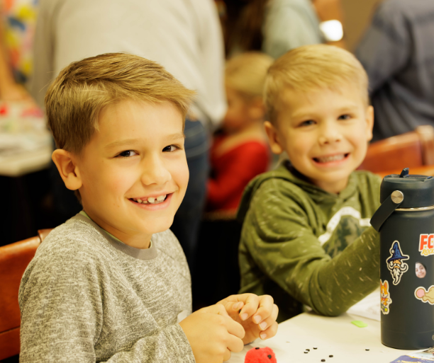 Two young boys with light-colored hair smiling at the camera, sitting at a table with a water bottle and small craft items in front of them, in a crowded indoor setting.