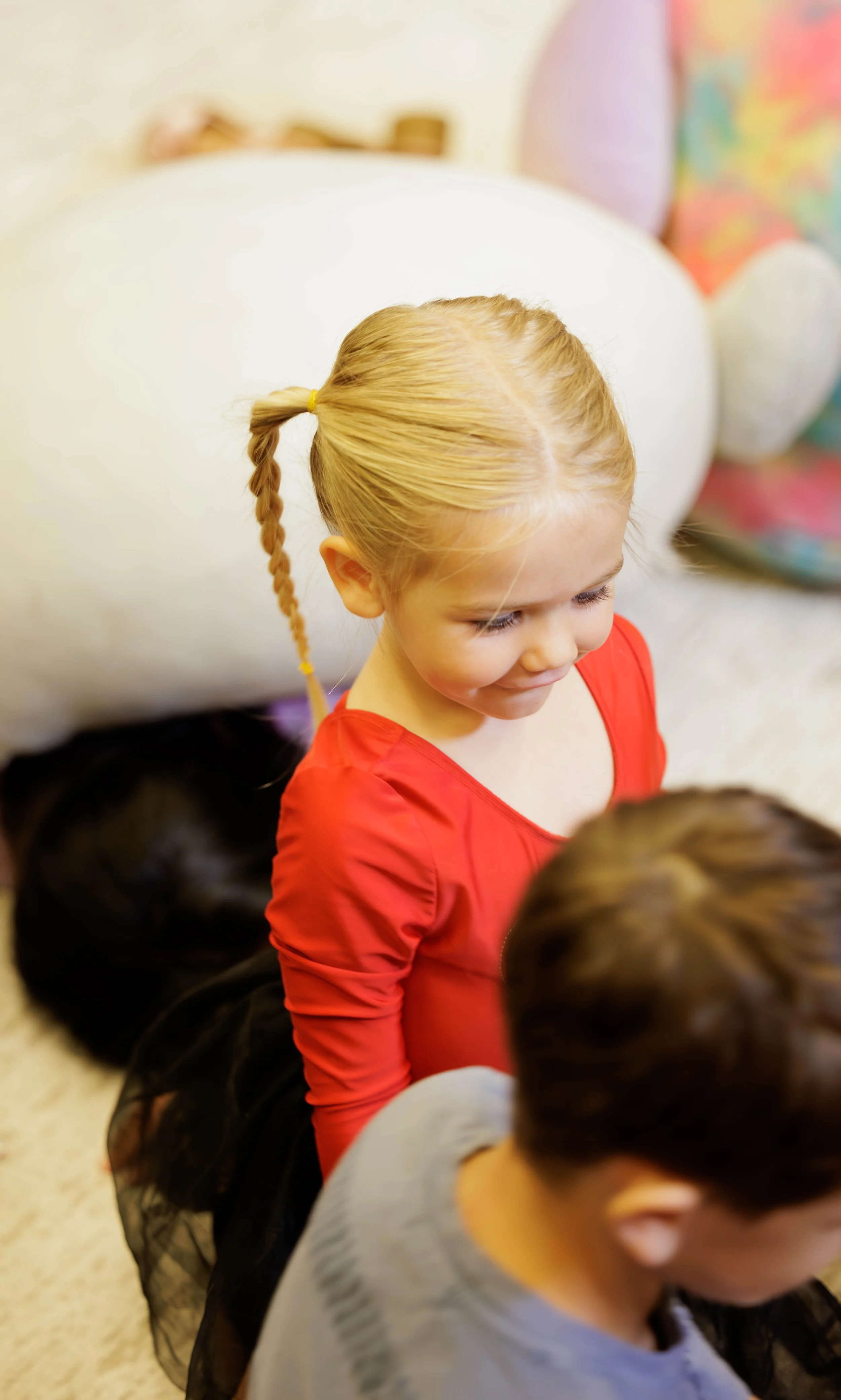 A young girl with blonde hair in a braid, wearing a red shirt, sitting on the floor with other children around her, in a colorful, indoor setting.
