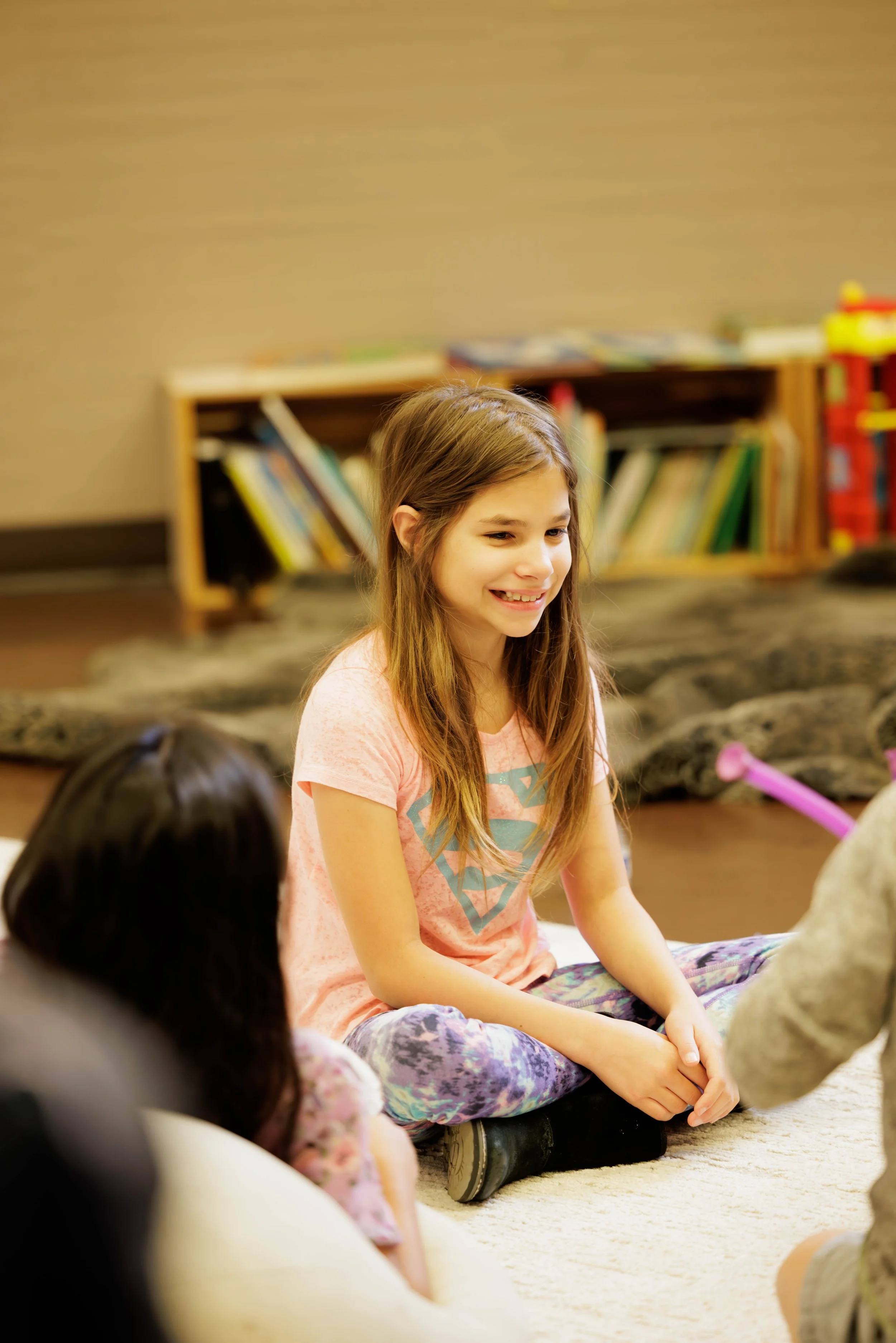 A girl with long brown hair sitting cross-legged on a beige carpet, smiling, wearing a pink Superman t-shirt and tie-dye leggings, engaged in a conversation with children in a room with a bookshelf and toys in the background.