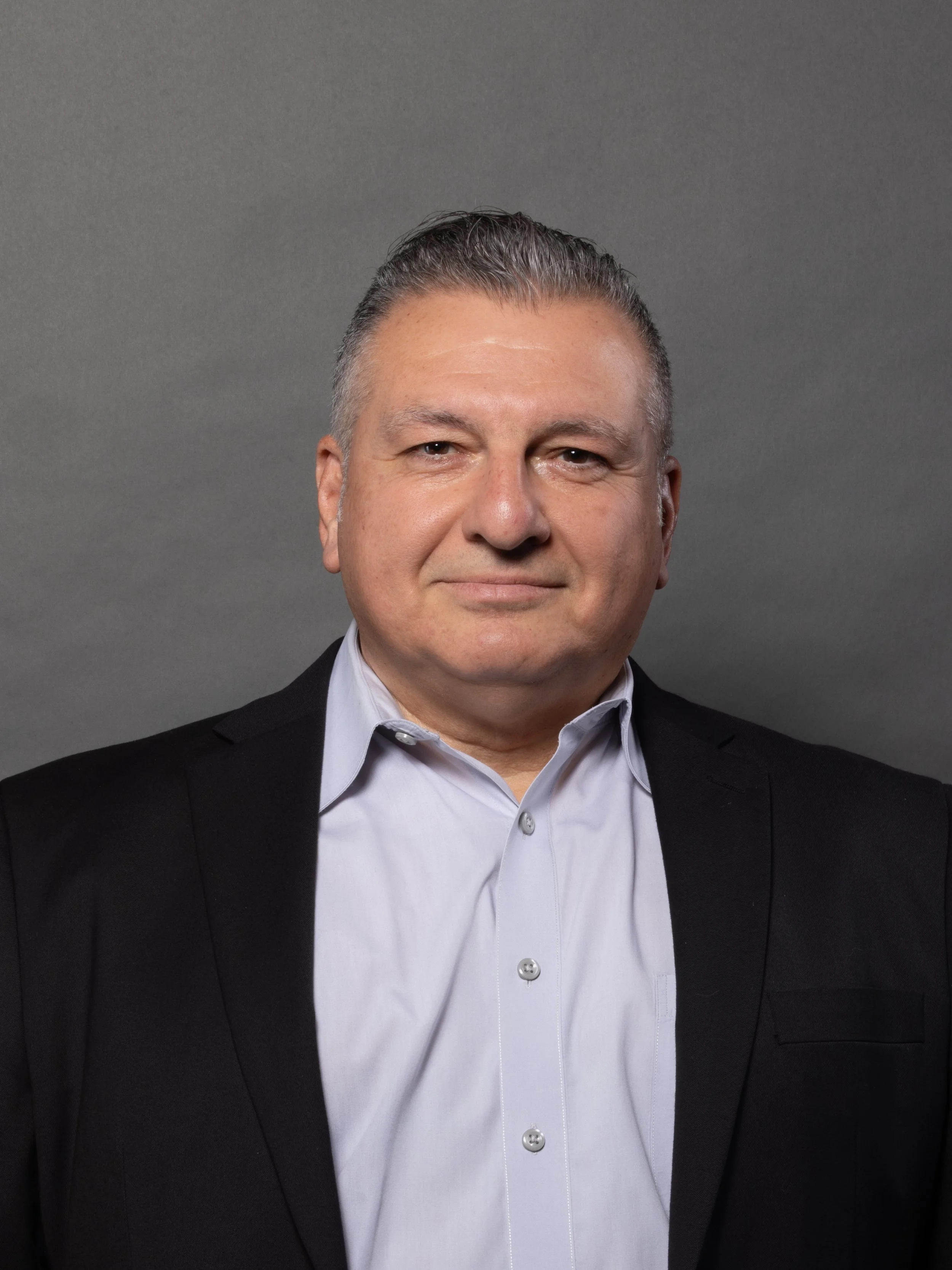 A middle-aged man with short gray hair, wearing a dark suit jacket and a light-colored dress shirt, is smiling softly against a gray background.