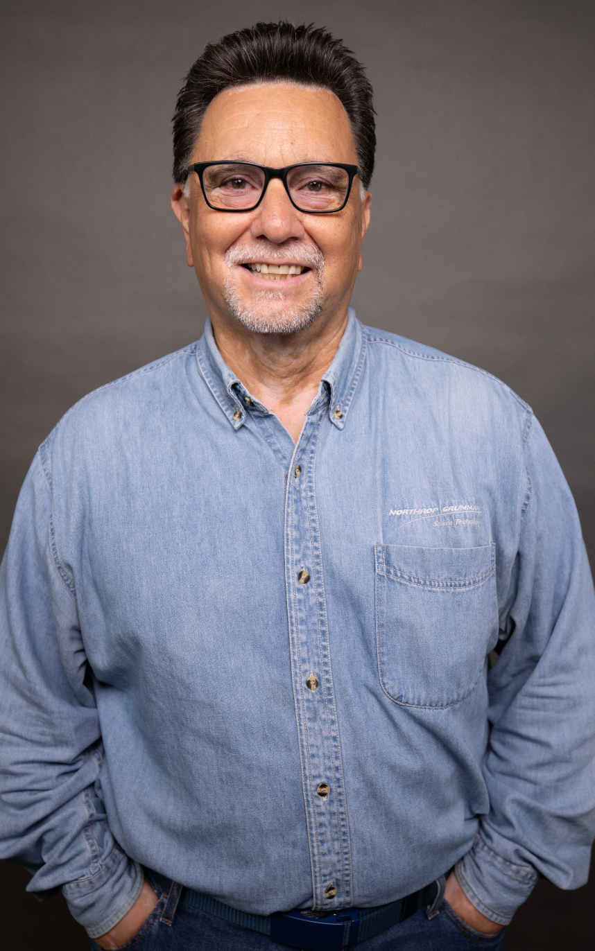 A middle-aged man with dark hair, glasses, and a goatee, smiling, wearing a light blue denim shirt against a gray background.