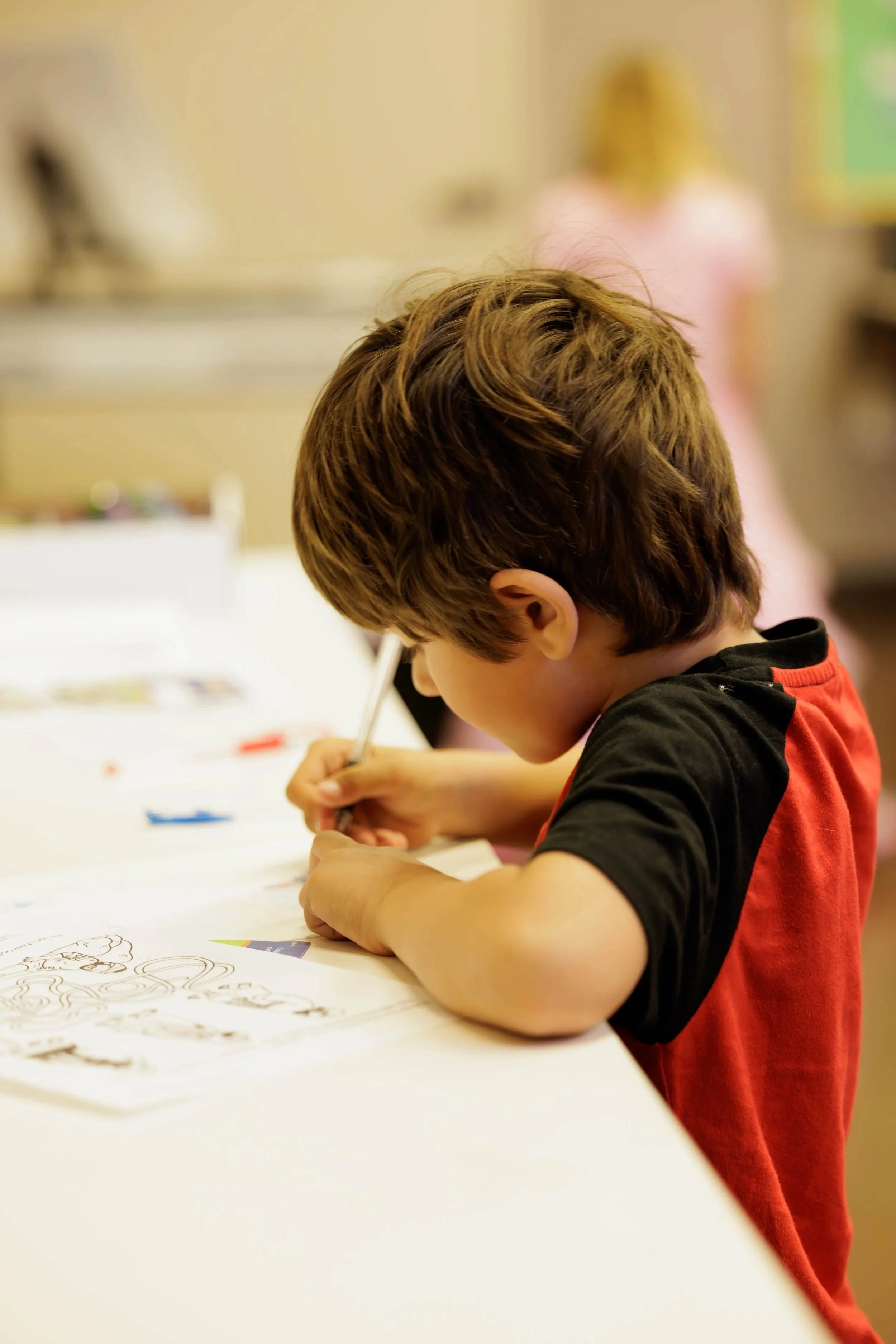 A young boy with brown hair, wearing a red and black shirt, is drawing or coloring on a piece of paper at a table. In the background, there is a woman with blonde hair working at a computer.