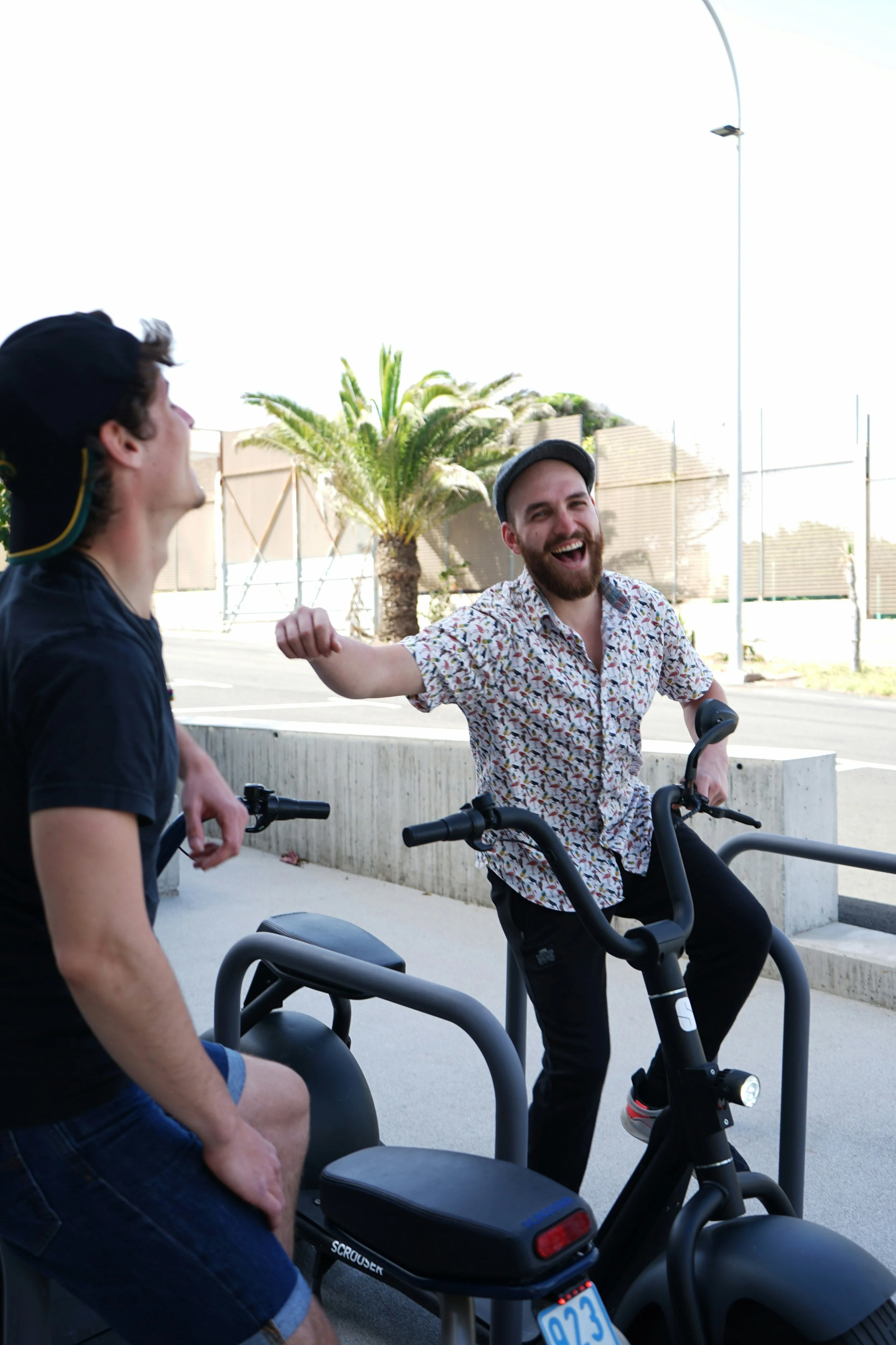 Two young men enjoying a fun moment; one sitting on a scooter and the other standing with a wide smile, on a sidewalk with palm trees and a fence in the background.