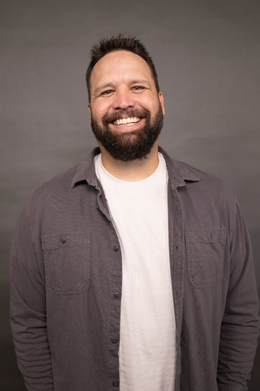 Portrait of a smiling man with a beard, wearing a white T-shirt and gray button-up shirt, against a plain gray background.