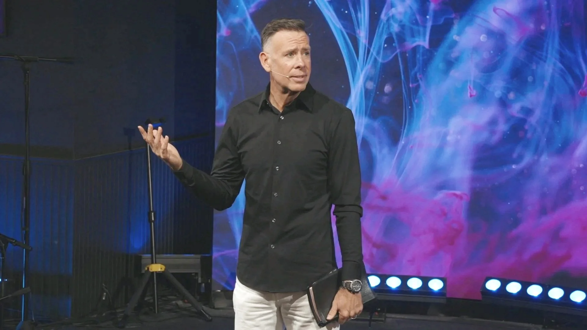 A man in a black shirt and white pants speaking on stage with a colorful abstract background.