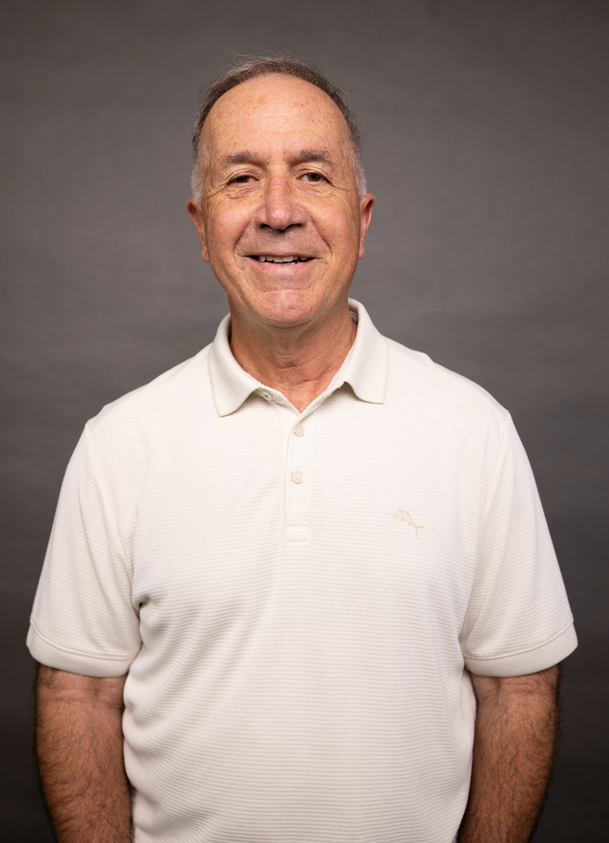 A middle-aged man with short hair and a friendly smile, wearing a white polo shirt, standing against a gray background.