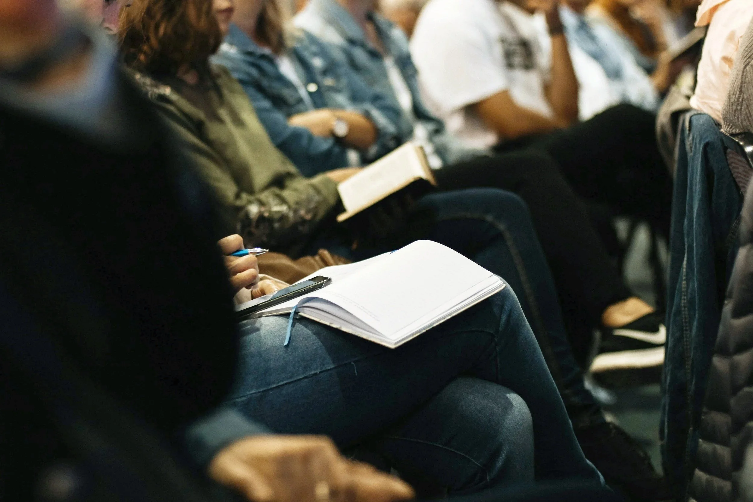 People attending a lecture or seminar, seated in chairs, some taking notes or reading.
