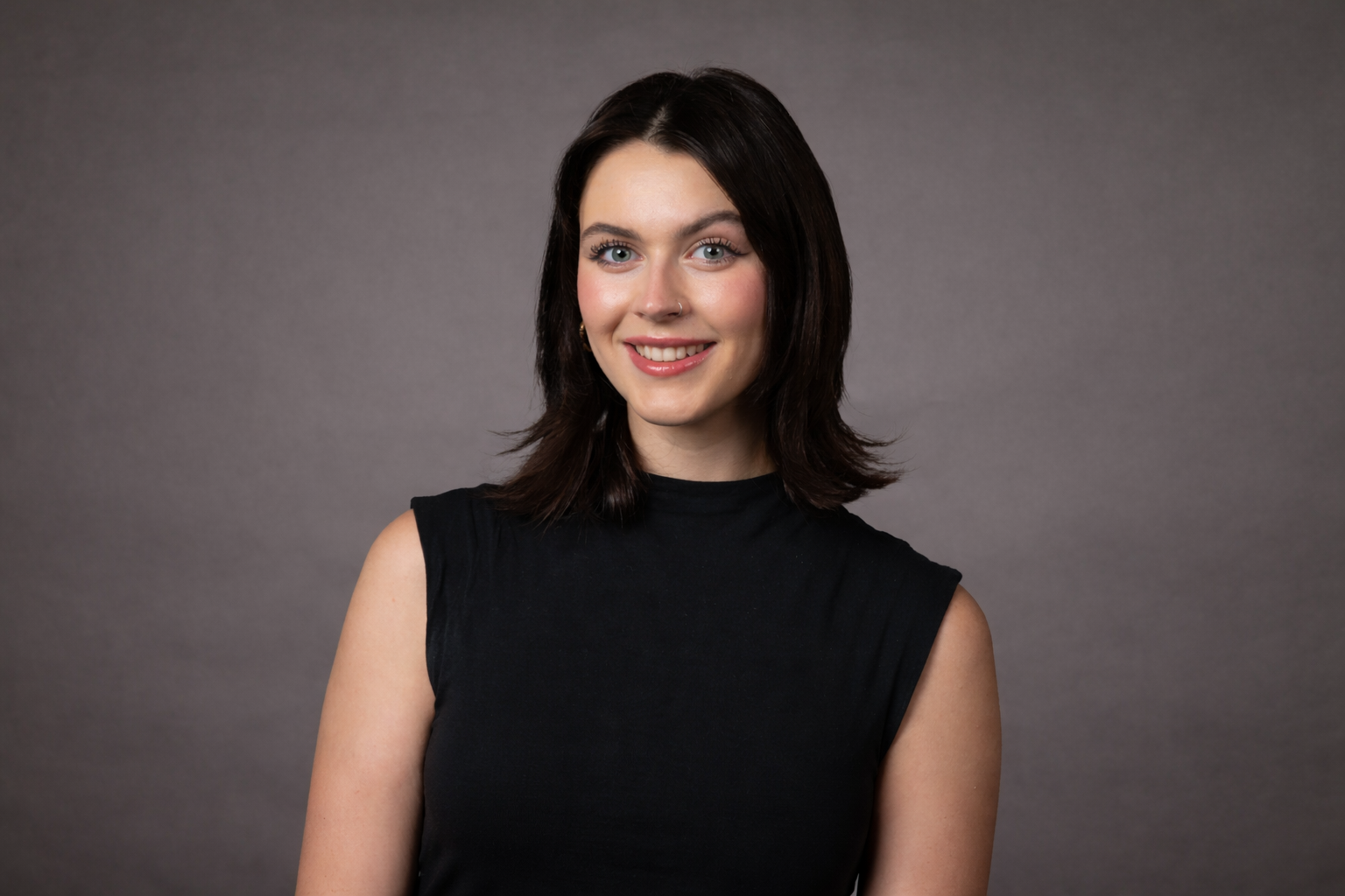 A young woman with dark brown hair and blue eyes, smiling, wearing a black sleeveless top, against a gray background.
