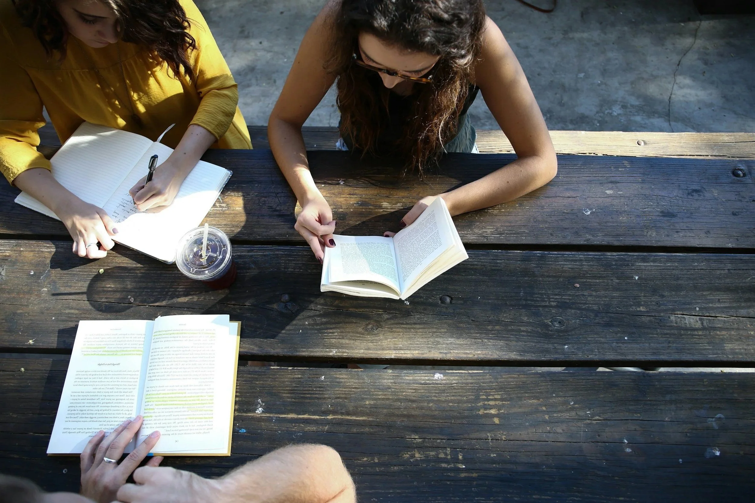 Overhead view of three people sitting at a wooden picnic table, reading books and drinking iced drinks with straws.