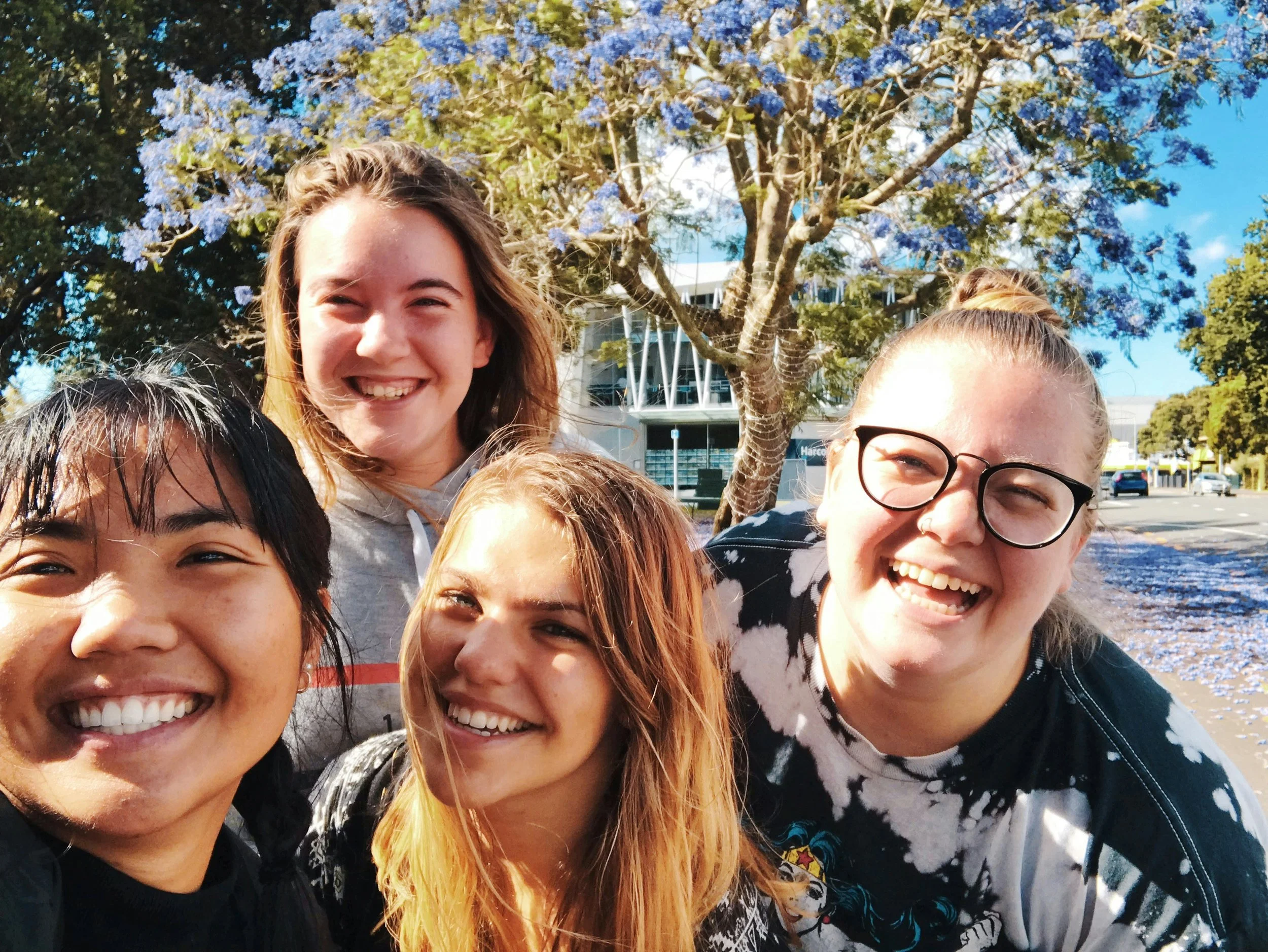 A group of five young women smiling outdoors with a purple flowering tree and a modern building in the background.