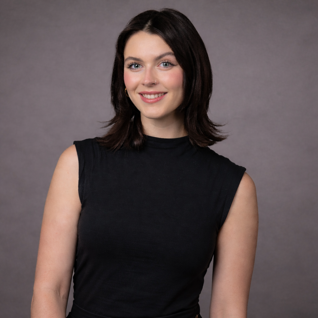 A young woman with dark hair, blue eyes, wearing a sleeveless black top, smiling in front of a plain gray background.