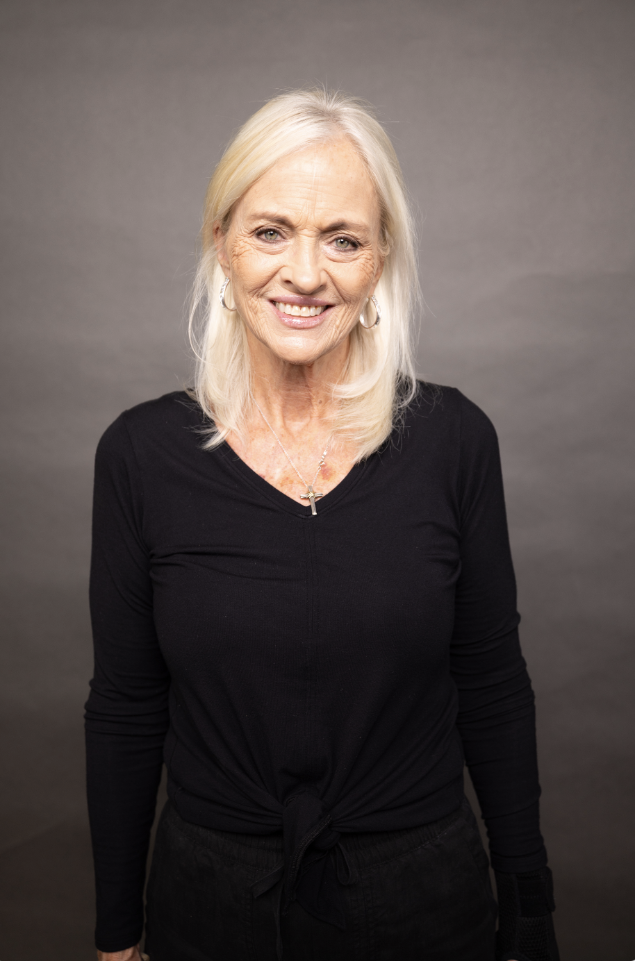 A portrait of an elderly woman with long white hair, smiling, wearing a black long sleeve top, silver hoop earrings, a necklace with a cross pendant, and standing against a plain gray background.