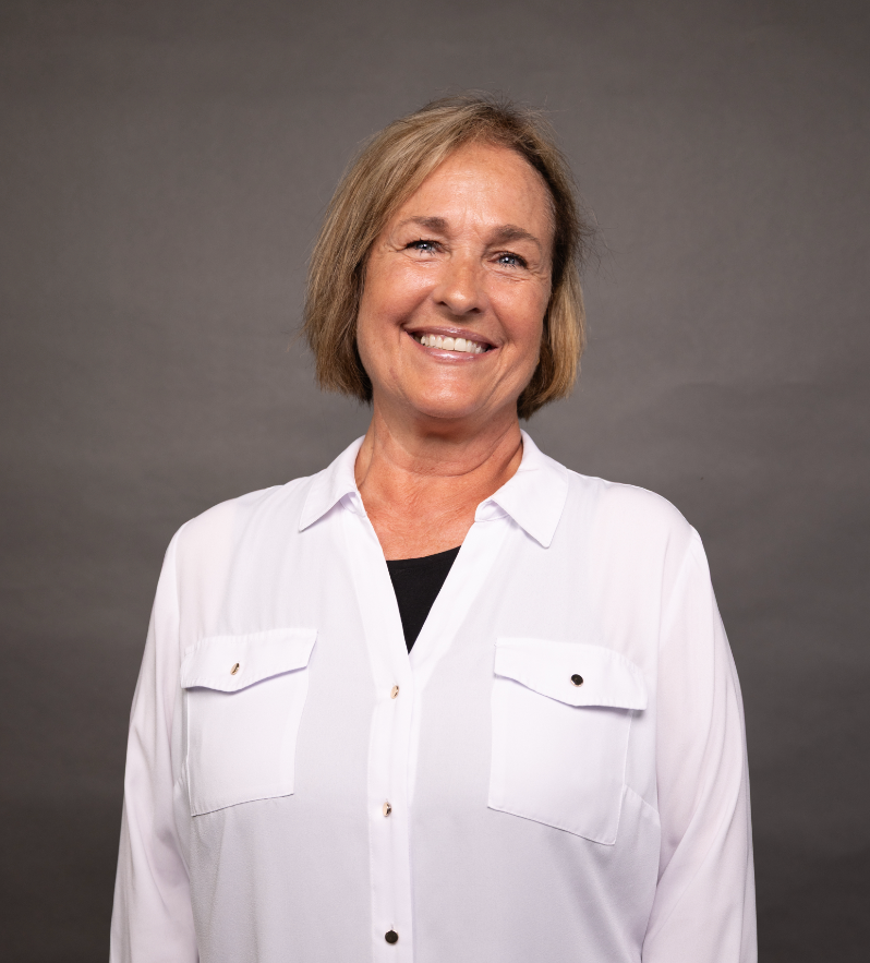 A smiling middle-aged woman with short blonde hair wearing a white button-up shirt and black undershirt against a gray background.