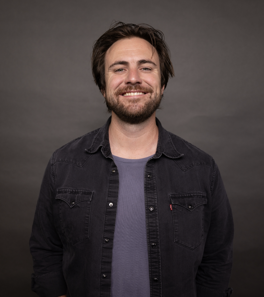 A smiling man with medium-length brown hair and a beard, wearing a dark denim shirt over a gray T-shirt, standing against a plain gray background.